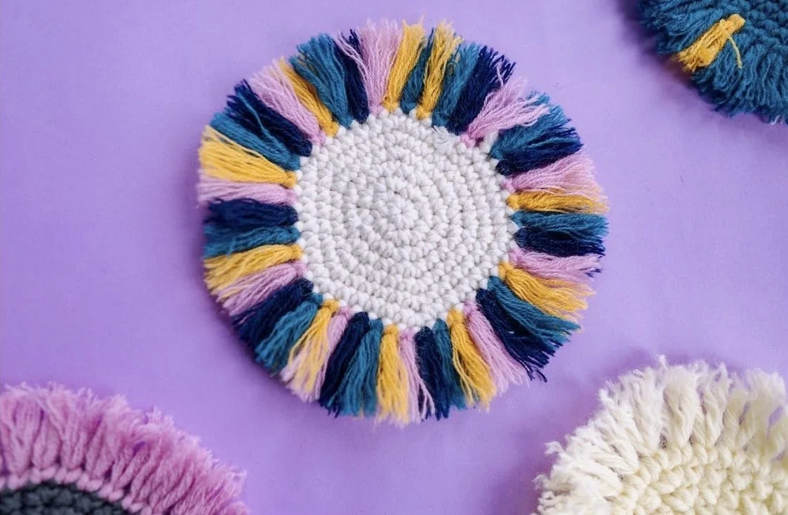 Colorful handmade round yarn coaster with a white center and multicolored fringe border, on a purple surface.