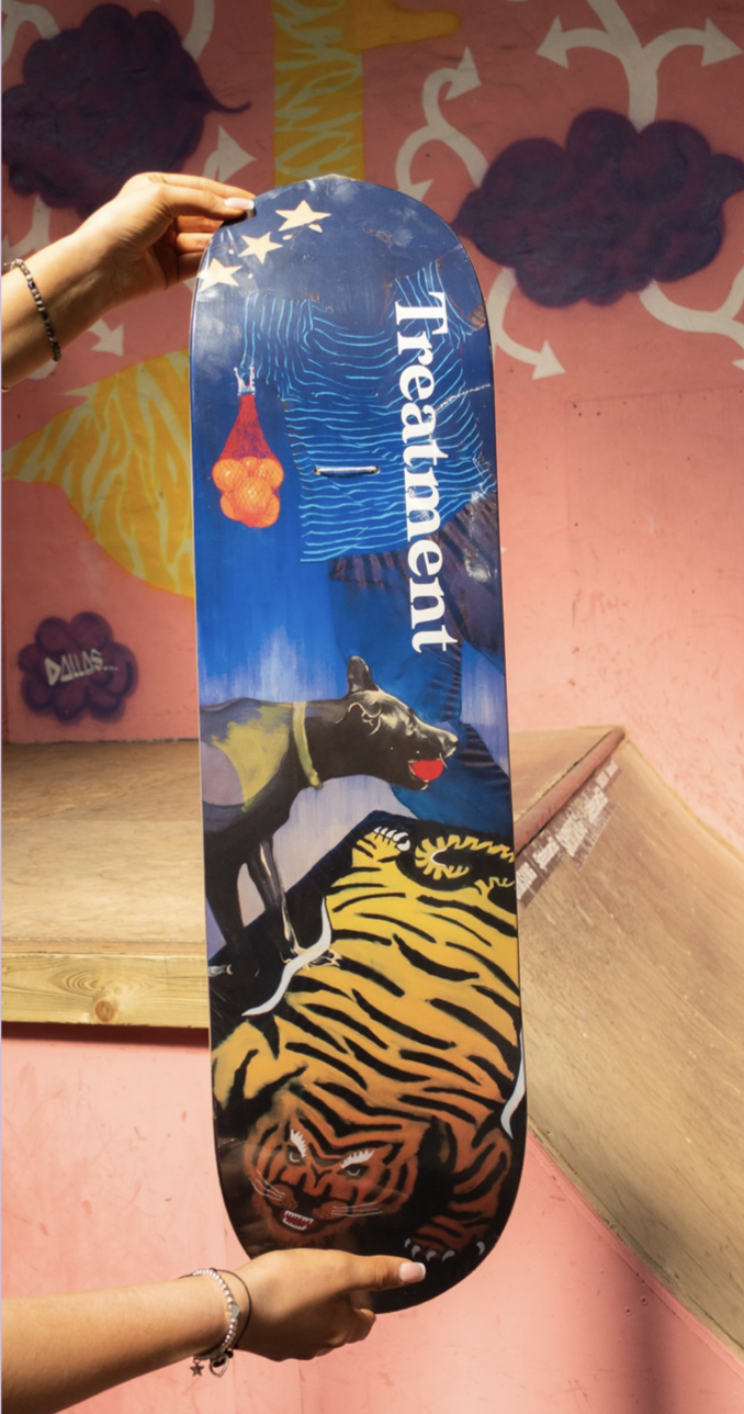 A skateboard deck with a colorful design featuring a black dog, a tiger, and the word 'Thetamer'. The background has illustrations of a cloud, a tree, and stars. The deck is held by two hands against a pink wall with painted clouds and trees.