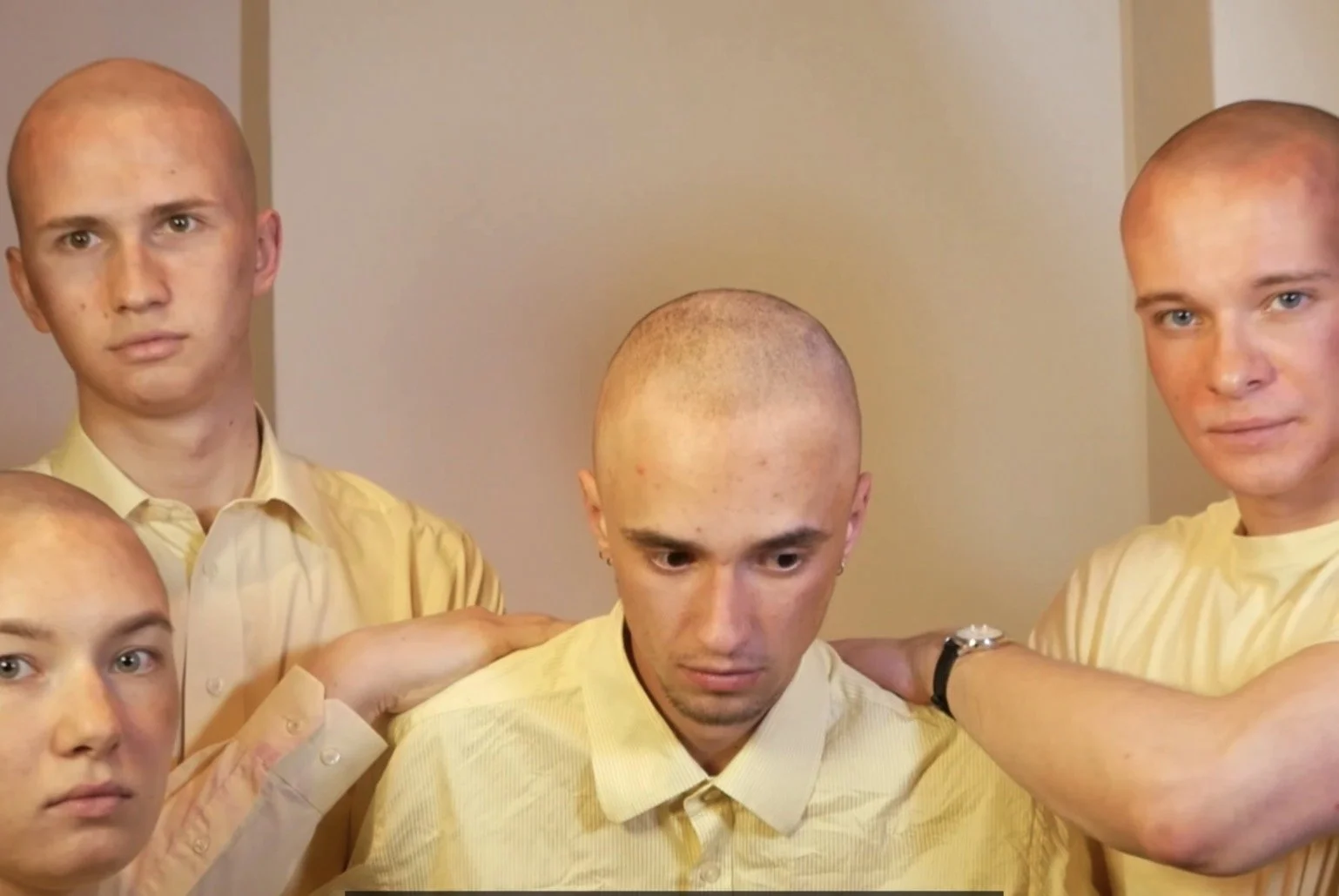 Five men with bald heads in yellow shirts, standing close together, with some placing their hands on the shoulders of others, in an indoor setting.