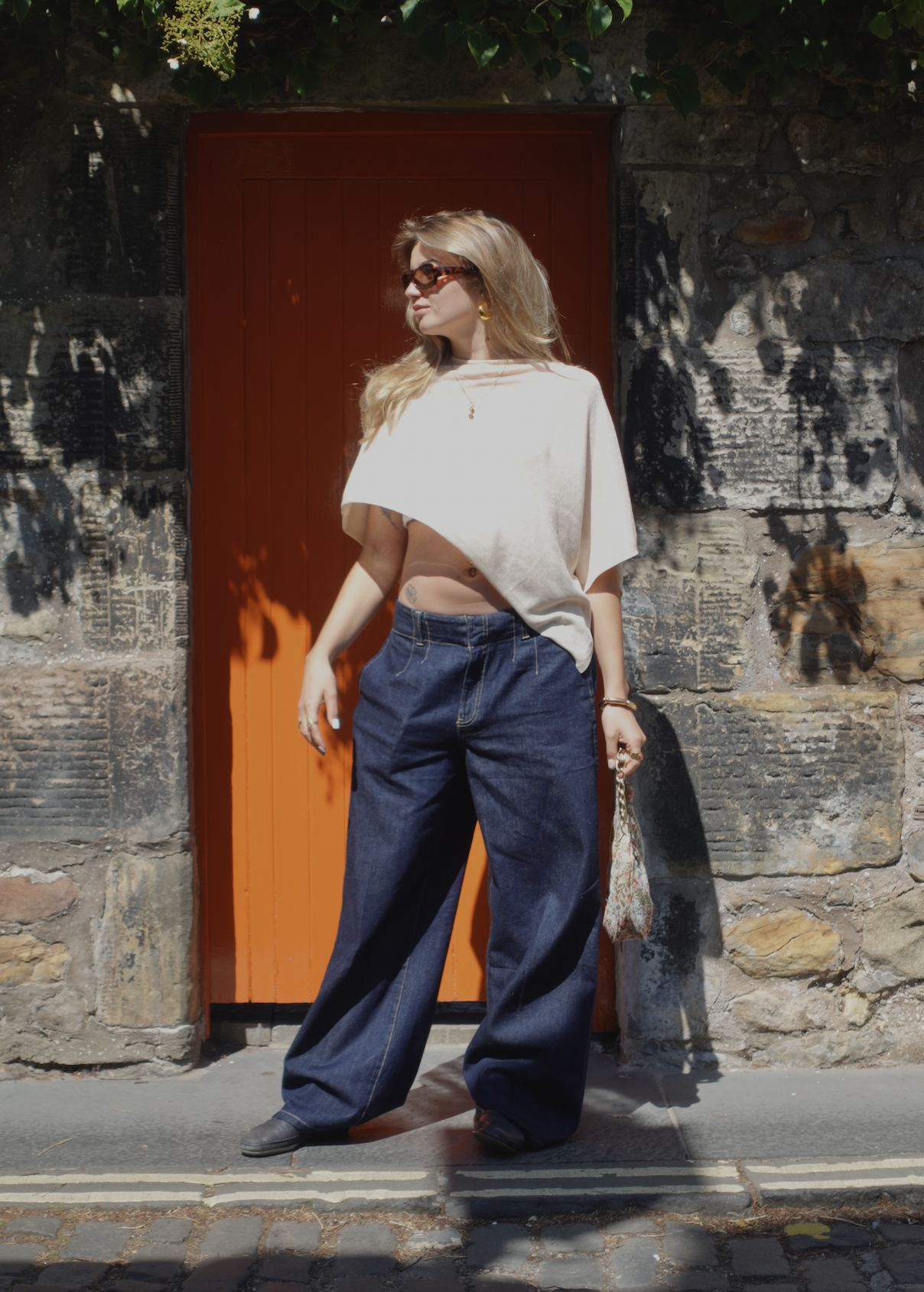 A woman with blonde hair and sunglasses standing in front of a red door and stone wall, wearing a beige crop top, loose dark blue jeans, and gold jewelry, holding a patterned bag.