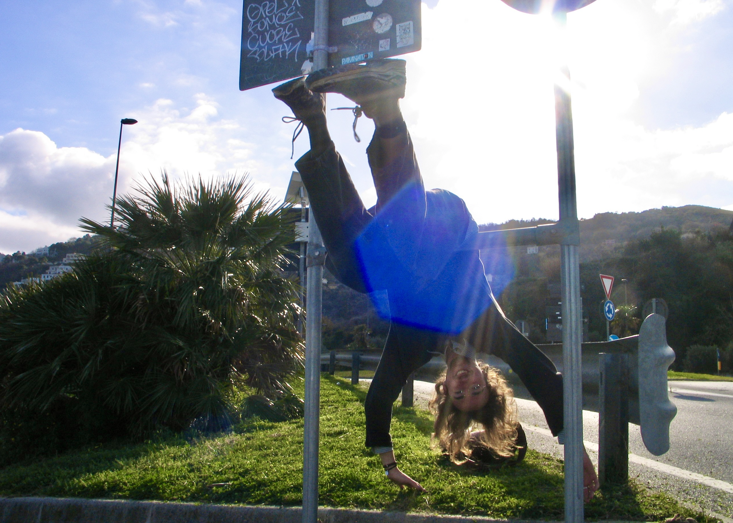 A young woman with glasses and curly hair doing a handstand under a basketball hoop, with her head close to the ground and legs extended upward. The photo is taken outside during the day with sunlight shining from the right side, partially obscuring the top of the hoop. There's greenery and a hill with houses in the background.