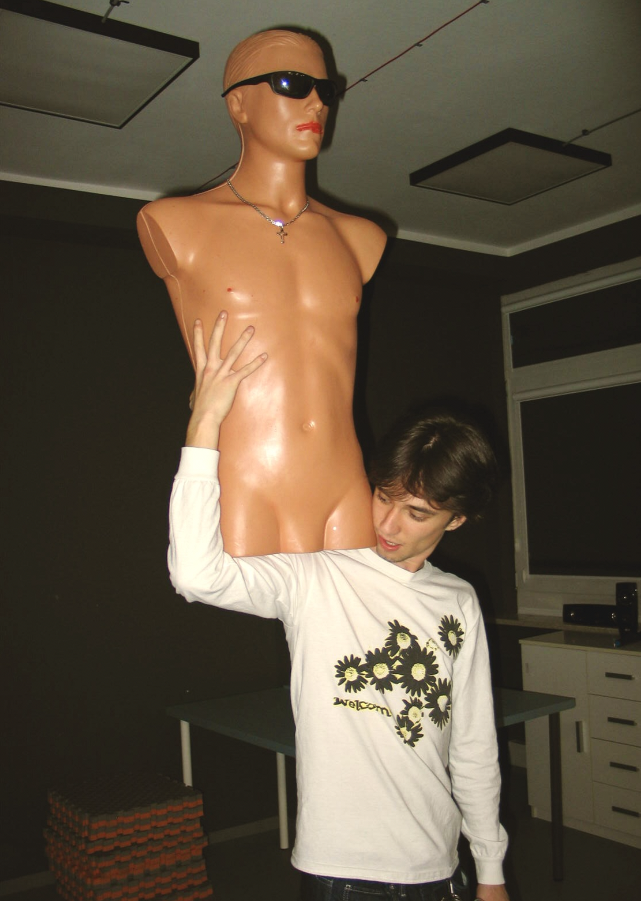 A young man is holding a life-sized mannequin head and torso with sunglasses and a necklace. The mannequin has a serious facial expression and is set against a plain background in a room with fluorescent ceiling lights.