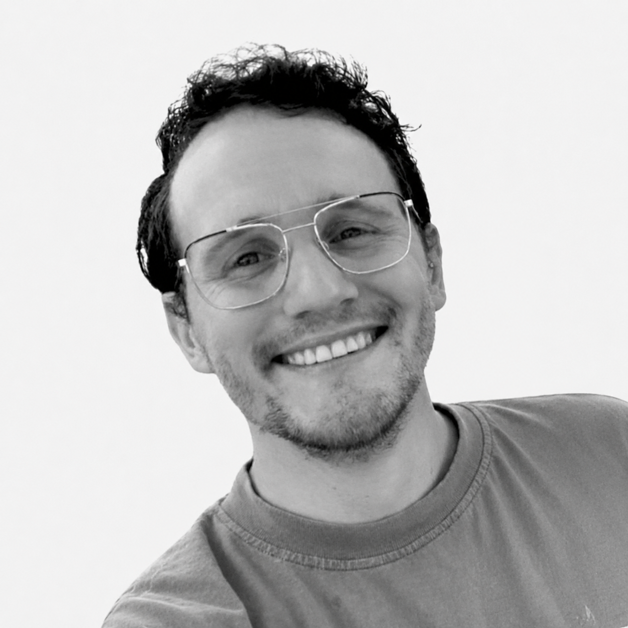 A smiling man with curly hair, glasses and a beard, wearing a t-shirt, against a plain light background.