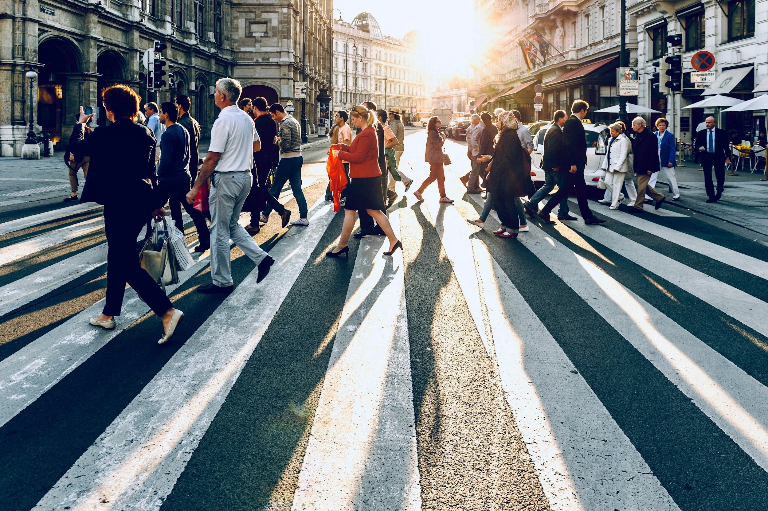 people crossing a wide city sidewalk with the sun low in the background