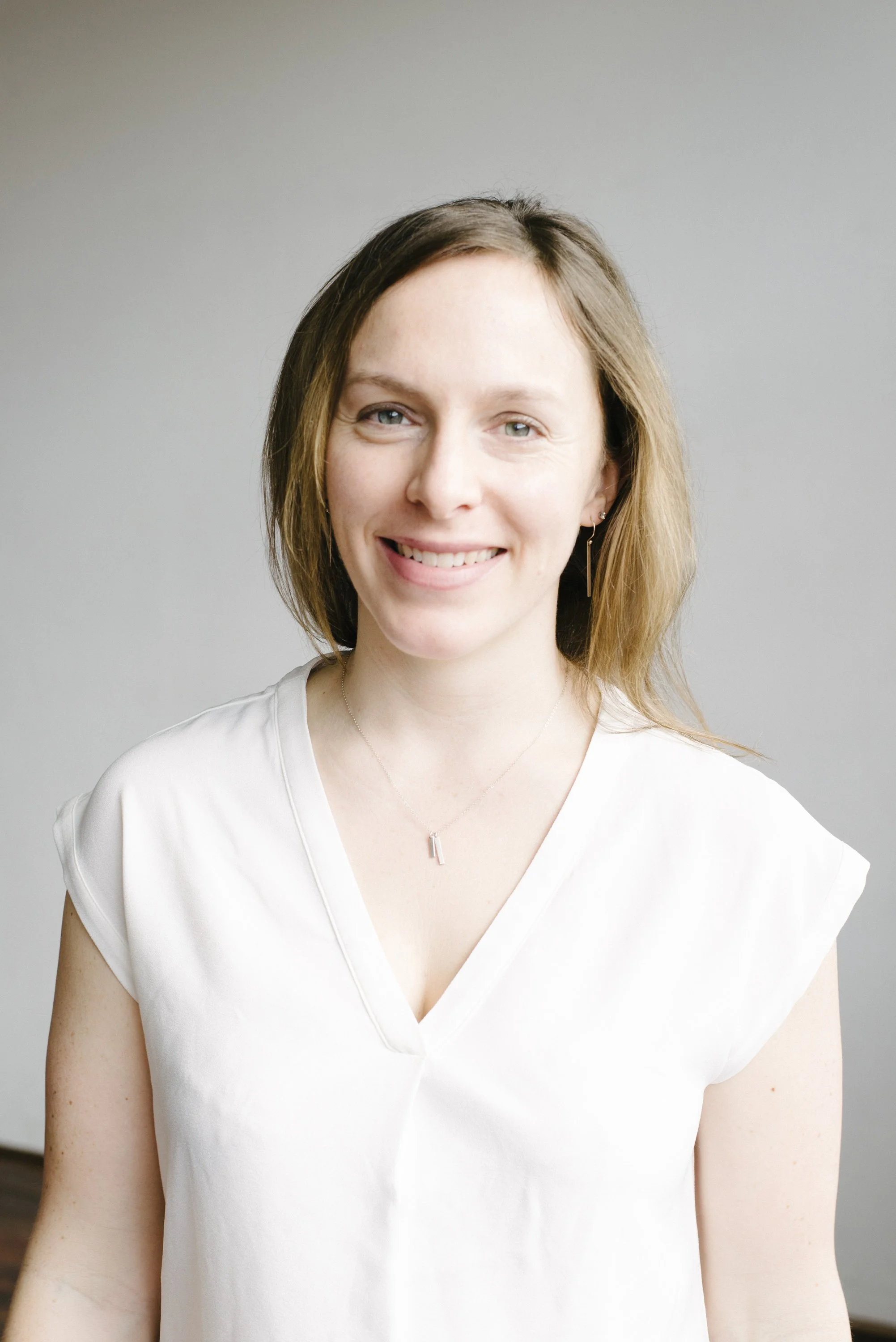 Portrait of a Rachel Drummond with shoulder-length light brown hair, smiling, wearing a white V-neck blouse, and a delicate necklace, standing against a plain gray background.