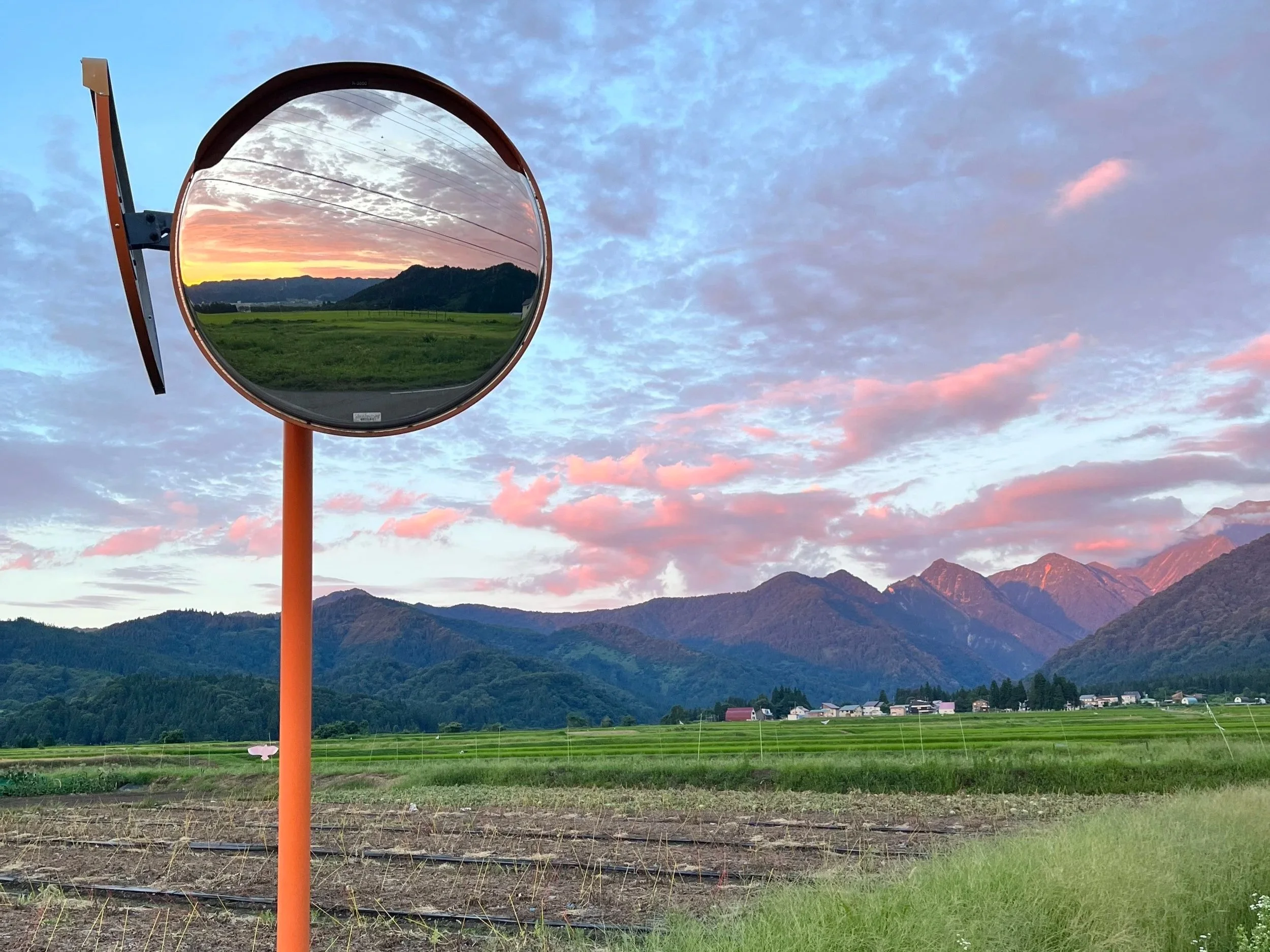 A convex traffic mirror reflecting mountains, a sunset sky with clouds, and a distant field with some houses.