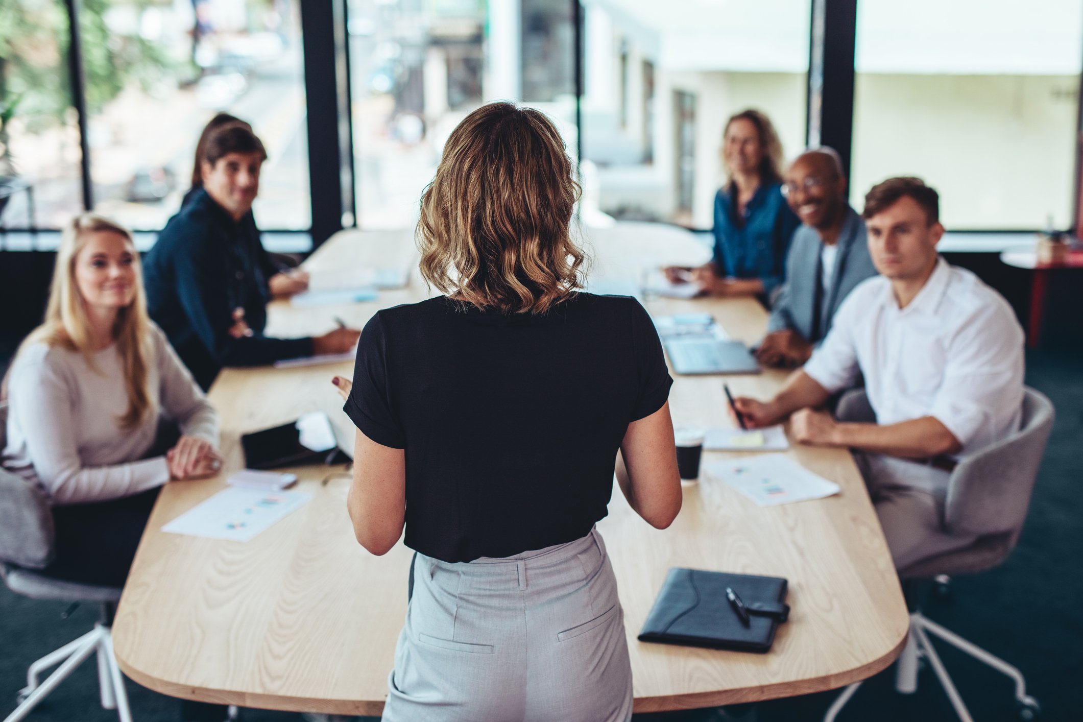 Eine Frau steht vor einem Konferenztisch und spricht zu einer Gruppe von fünf Menschen, die um den Tisch sitzen. Die Gruppe besteht aus Männern und Frauen, die Notizen und elektronische Geräte vor sich haben, in einem modernen Büro mit großen Fenstern im Hintergrund.