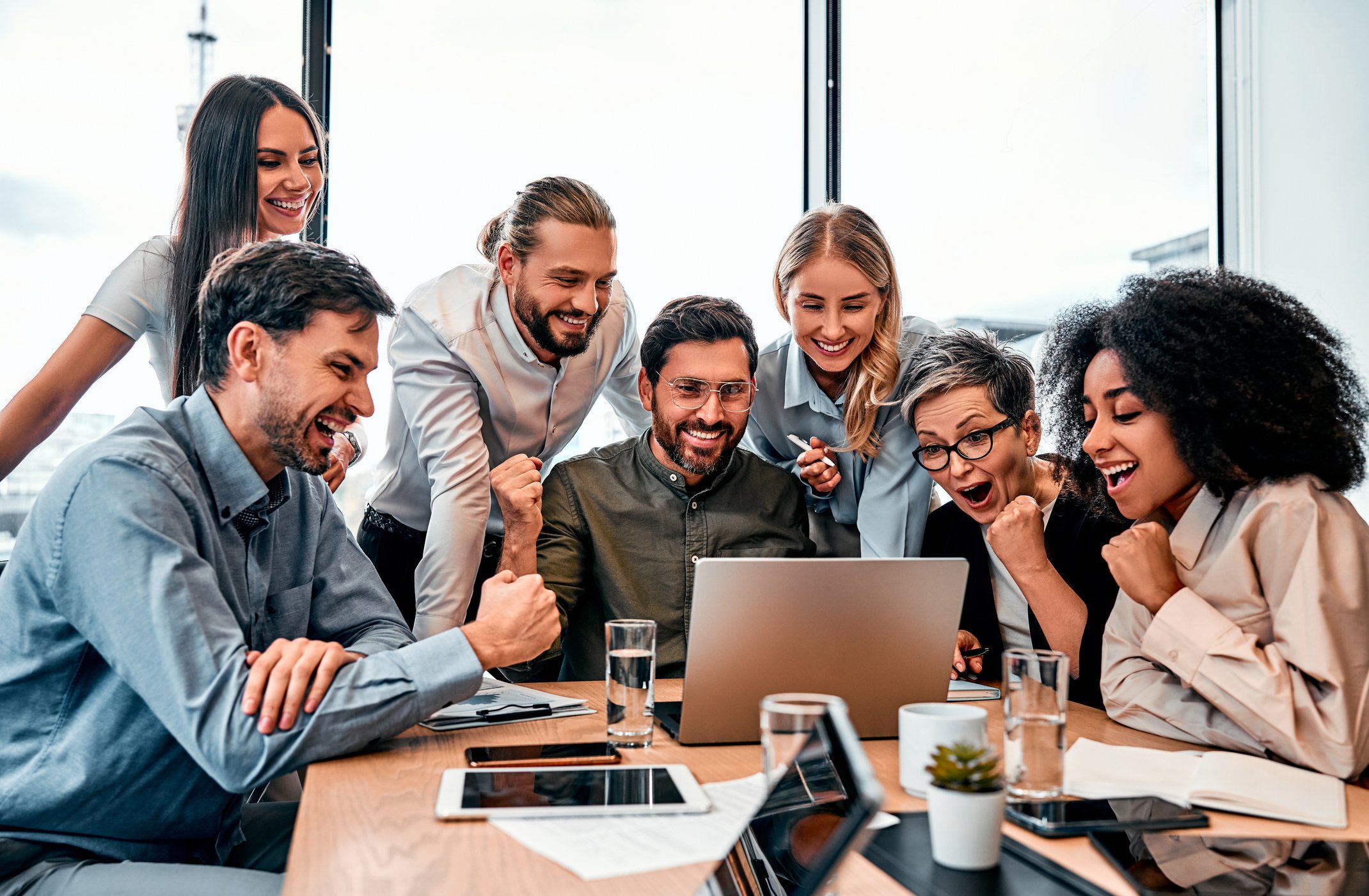 Eine Gruppe von sechs Menschen in einem Büro um einen Laptop, sie lachen und jubeln gemeinsam.