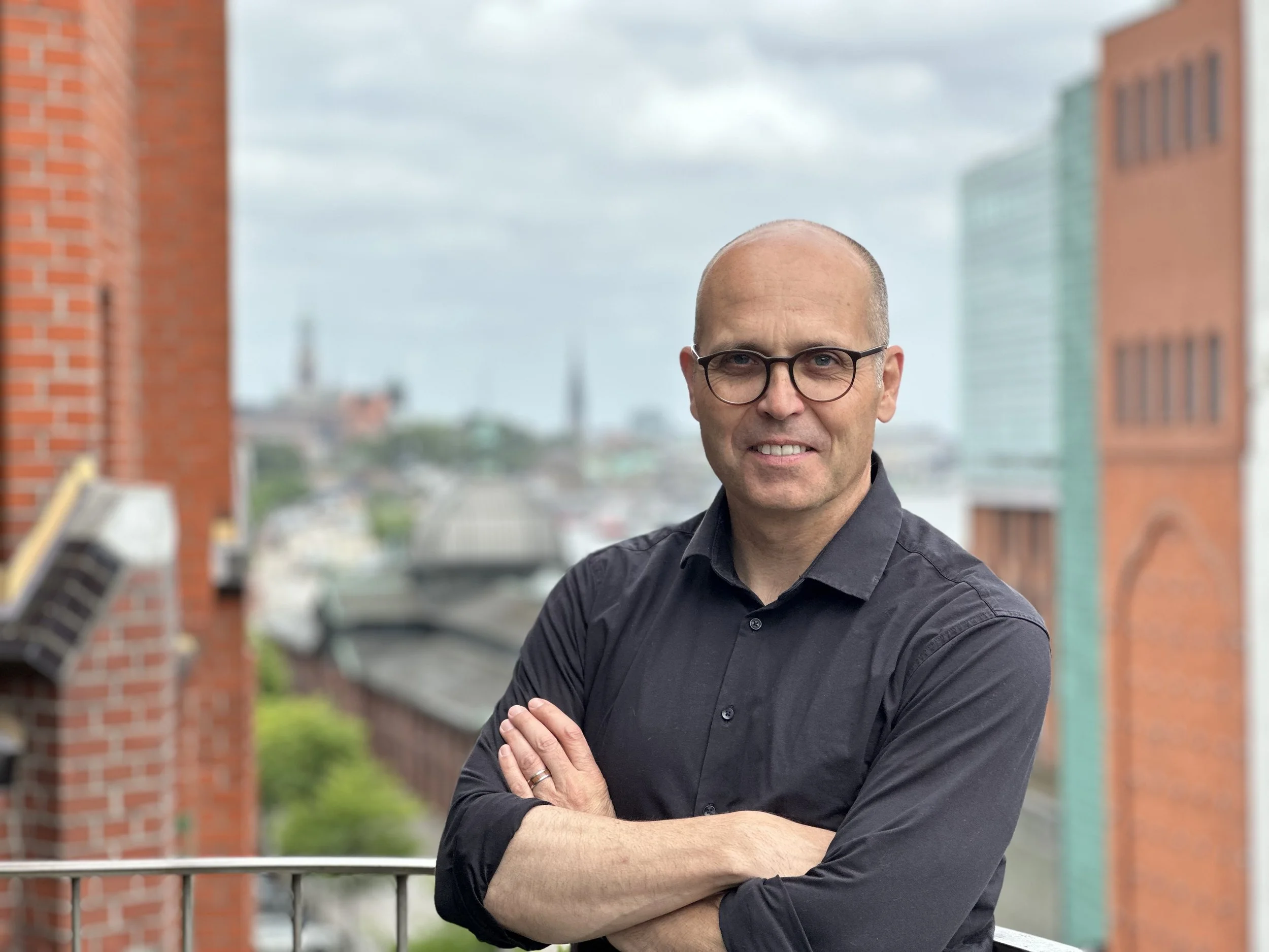 Ein Mann trägt eine schwarze Hemd und Brille, steht auf einem Balkon mit Blick auf die Stadt, hinter ihm sind Wolken am Himmel.