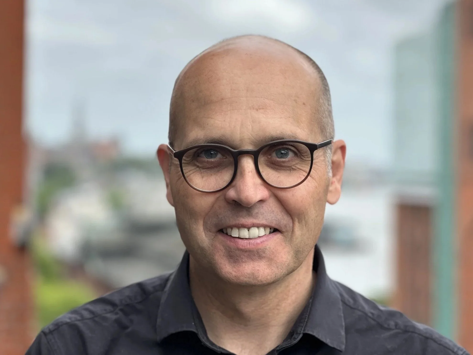 Ein Mann mit Brille und Glatze lächelt. Im Hintergrund ist eine verschwommene helle Umgebung zu sehen.