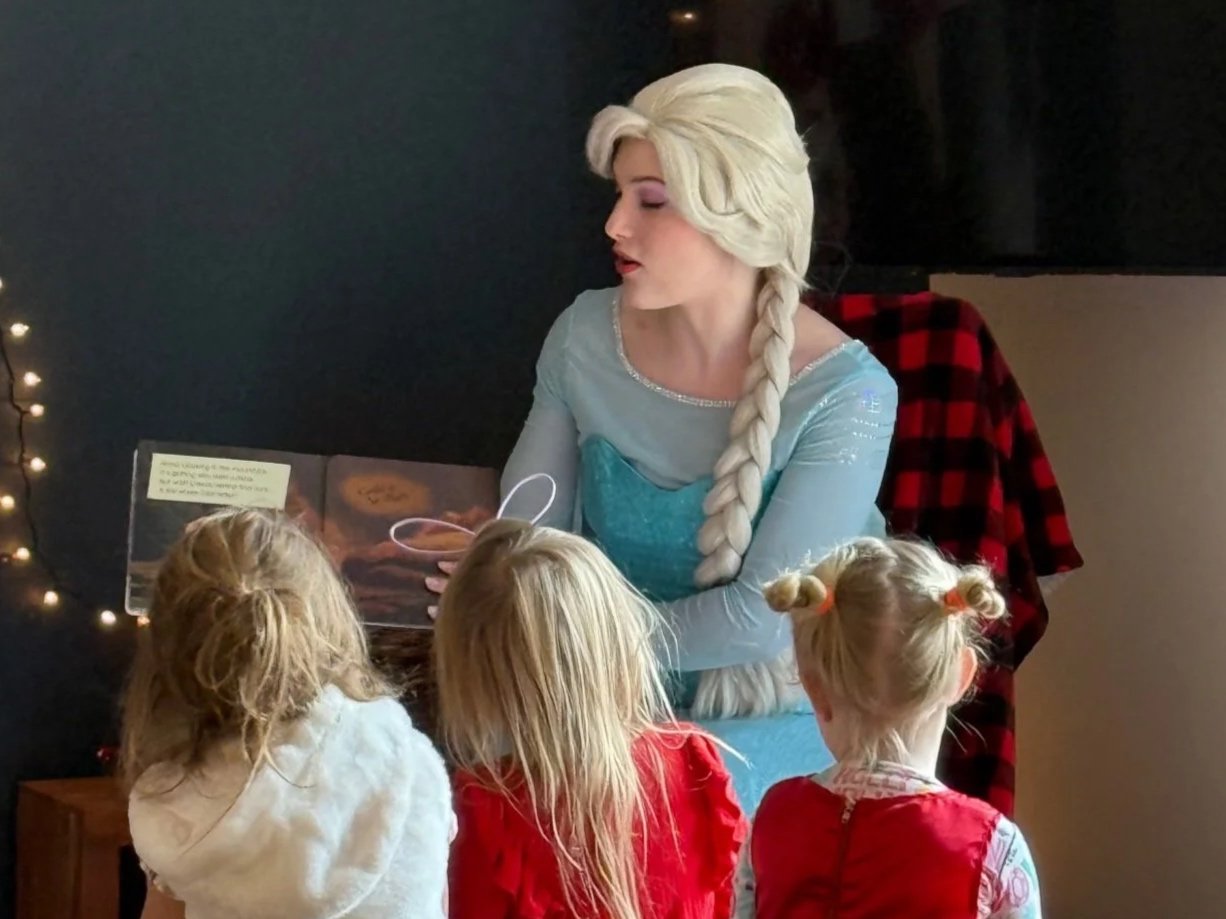 Elsa, from Frozen, reading a story to three young girls in a cozy room decorated with string lights.
