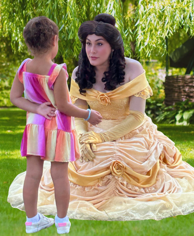 A woman dressed as Belle from Disney's 'Beauty and the Beast' kneeling on the grass, talking to a young girl in a colorful dress outdoors with green foliage in the background.
