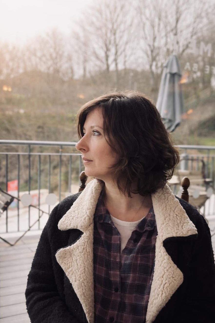 A woman with dark wavy hair looking to her left on a balcony with trees in the background during sunset.