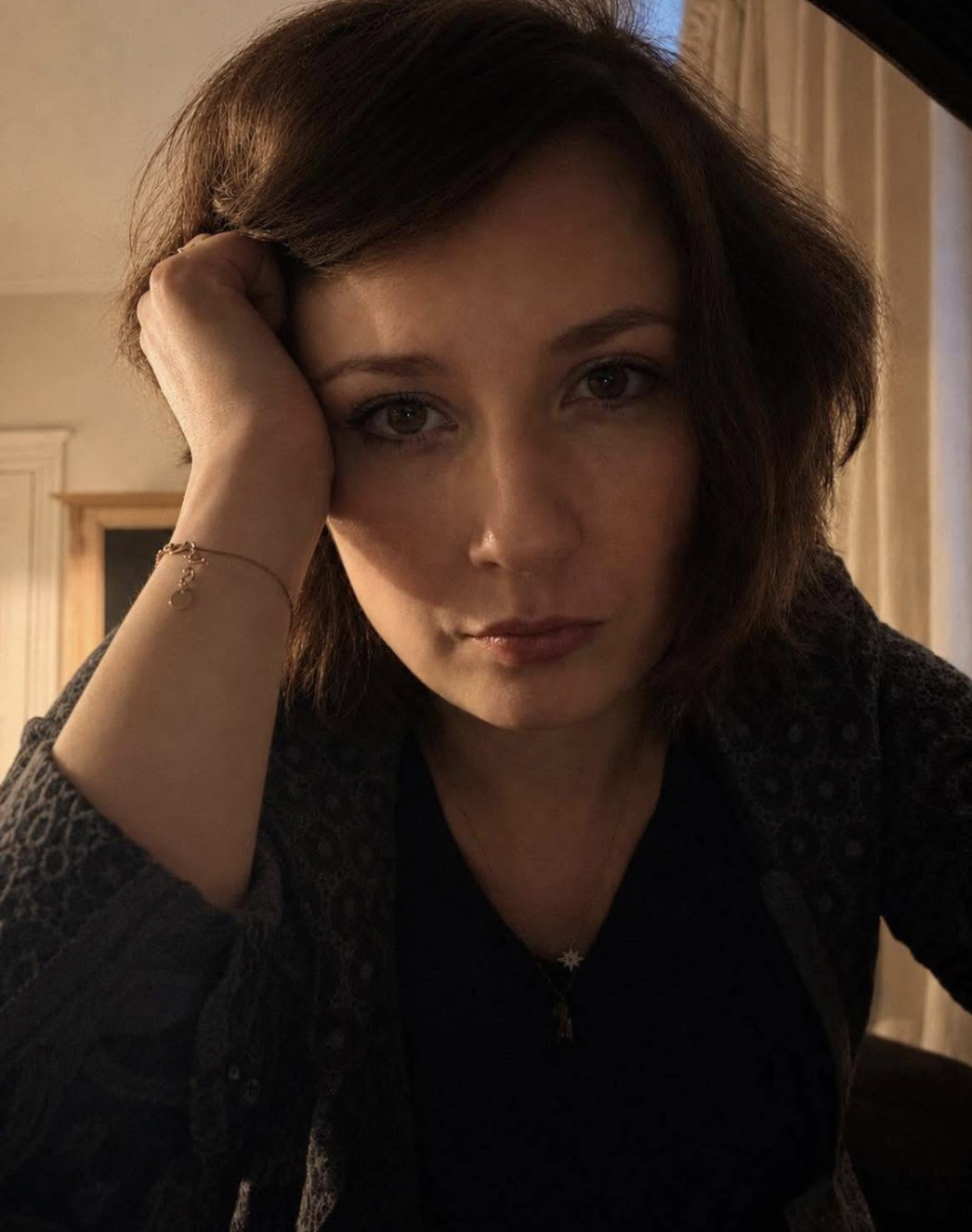 A woman with brown hair resting her head on her hand, wearing a dark top, a gold bracelet, and a star-shaped necklace, in a warmly lit room with wooden paneling.