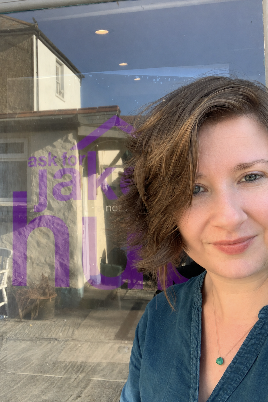 A woman with short brown hair and blue eyes standing outside, reflected in a glass window with purple text that reads 'ask for jake'.