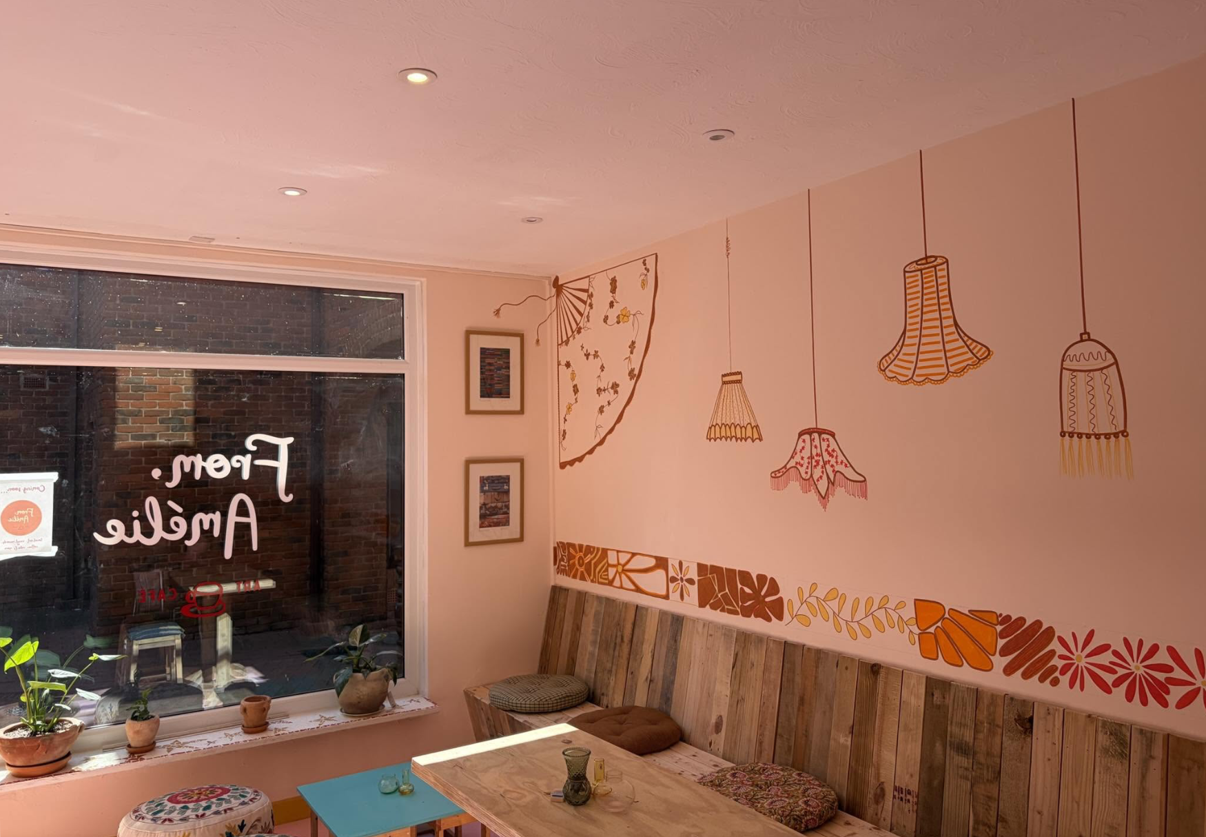 Interior of a cozy cafe with a wooden bench seat, a small wooden table adorned with candles, plants on the window sill, and framed pictures on the wall. The wall features decorative illustrations of hanging lamps and a floral corner. The large window shows a brick exterior.
