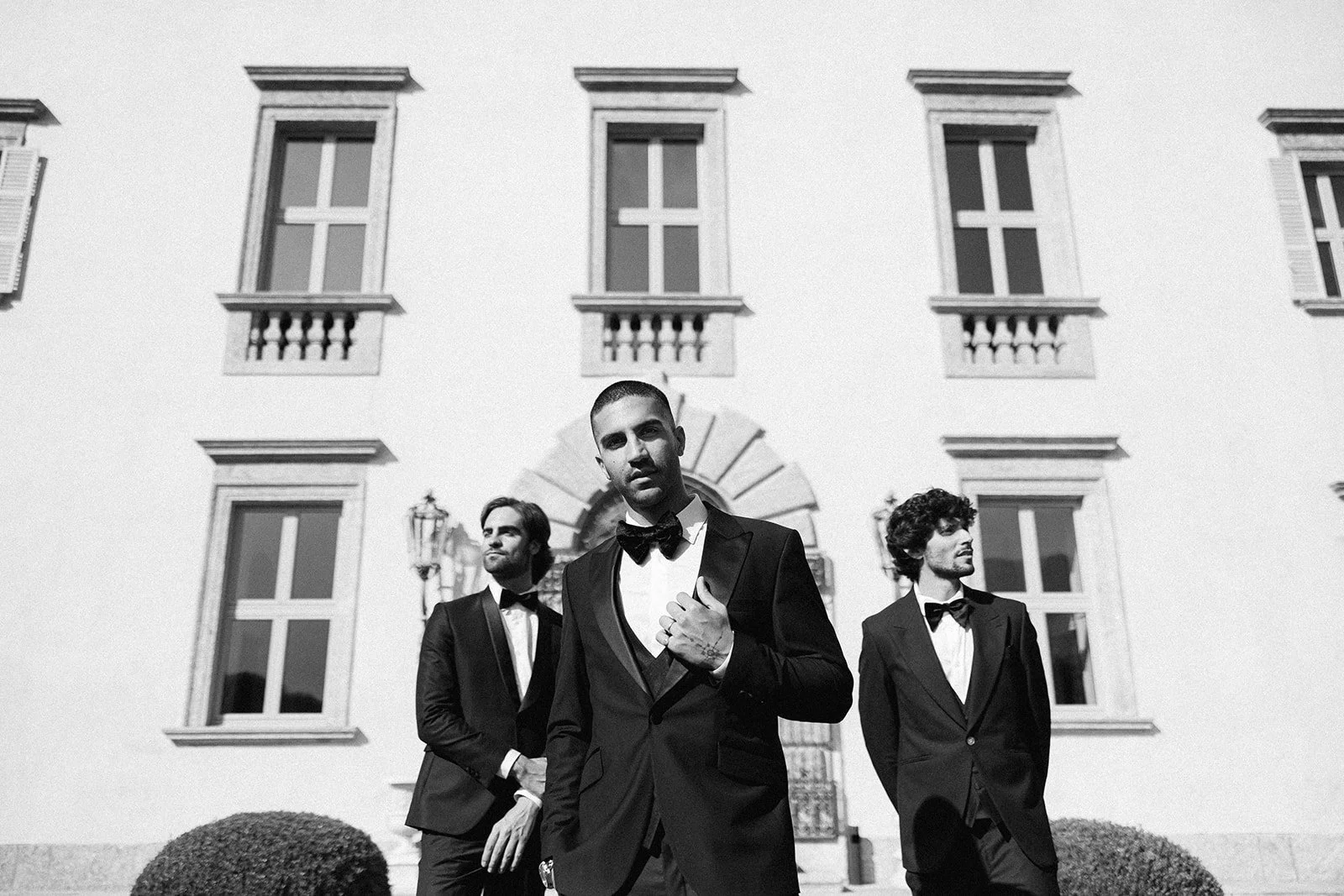 Three men dressed in tuxedos and bow ties standing outside in front of a large building with multiple windows and decorative shutters, black and white photograph.