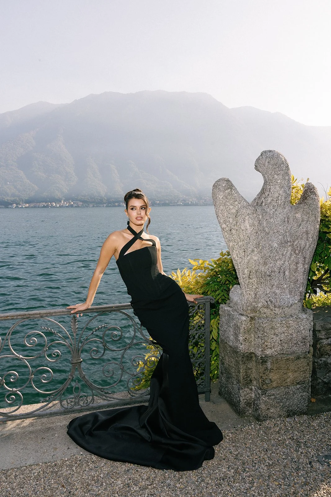 A woman in a black evening gown leaning on a decorative metal railing by a lake, with mountains in the background and a large stone sculpture of a hand nearby.