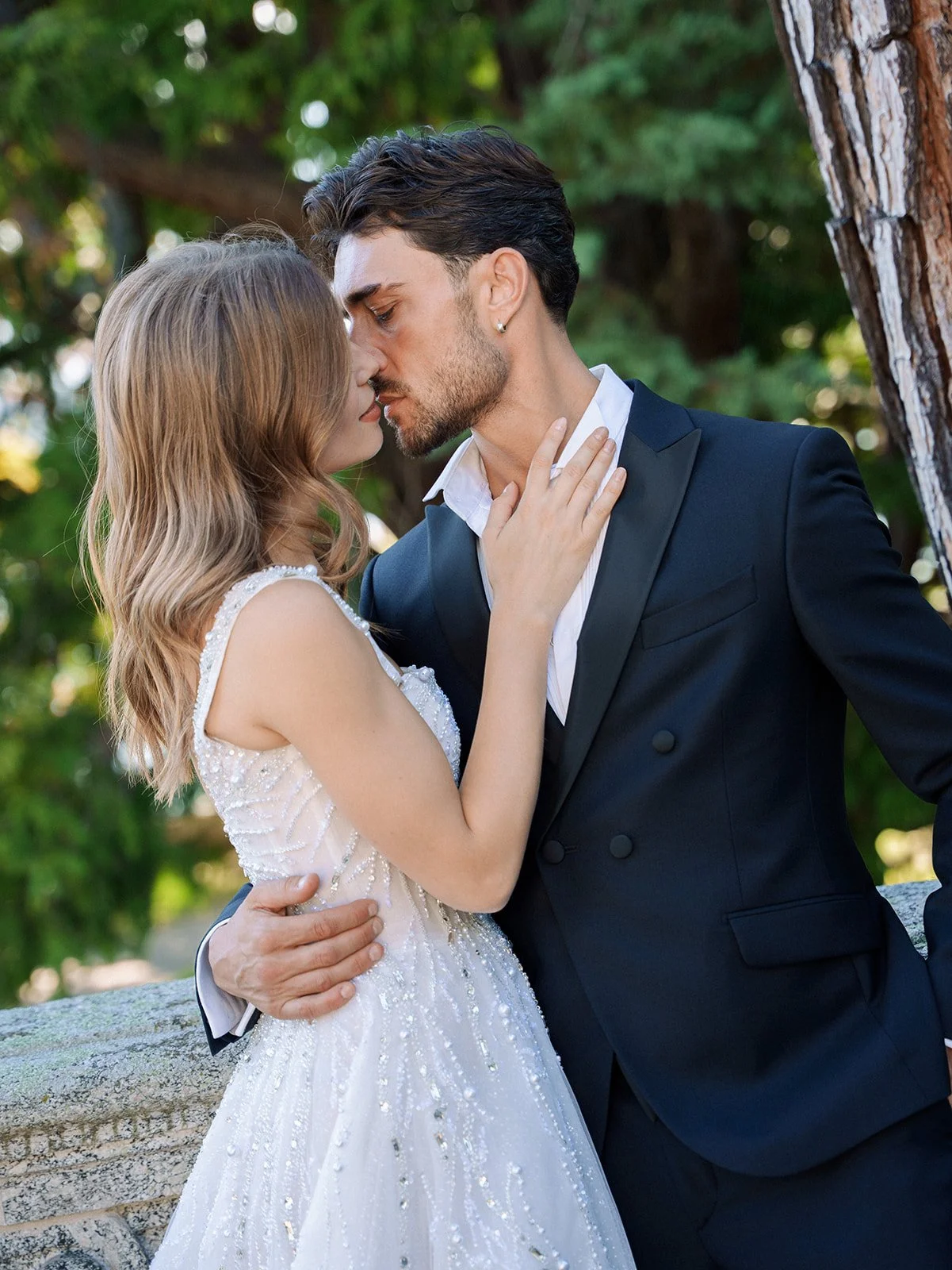 A bride and groom sharing a kiss outdoors in a lush green environment with trees.