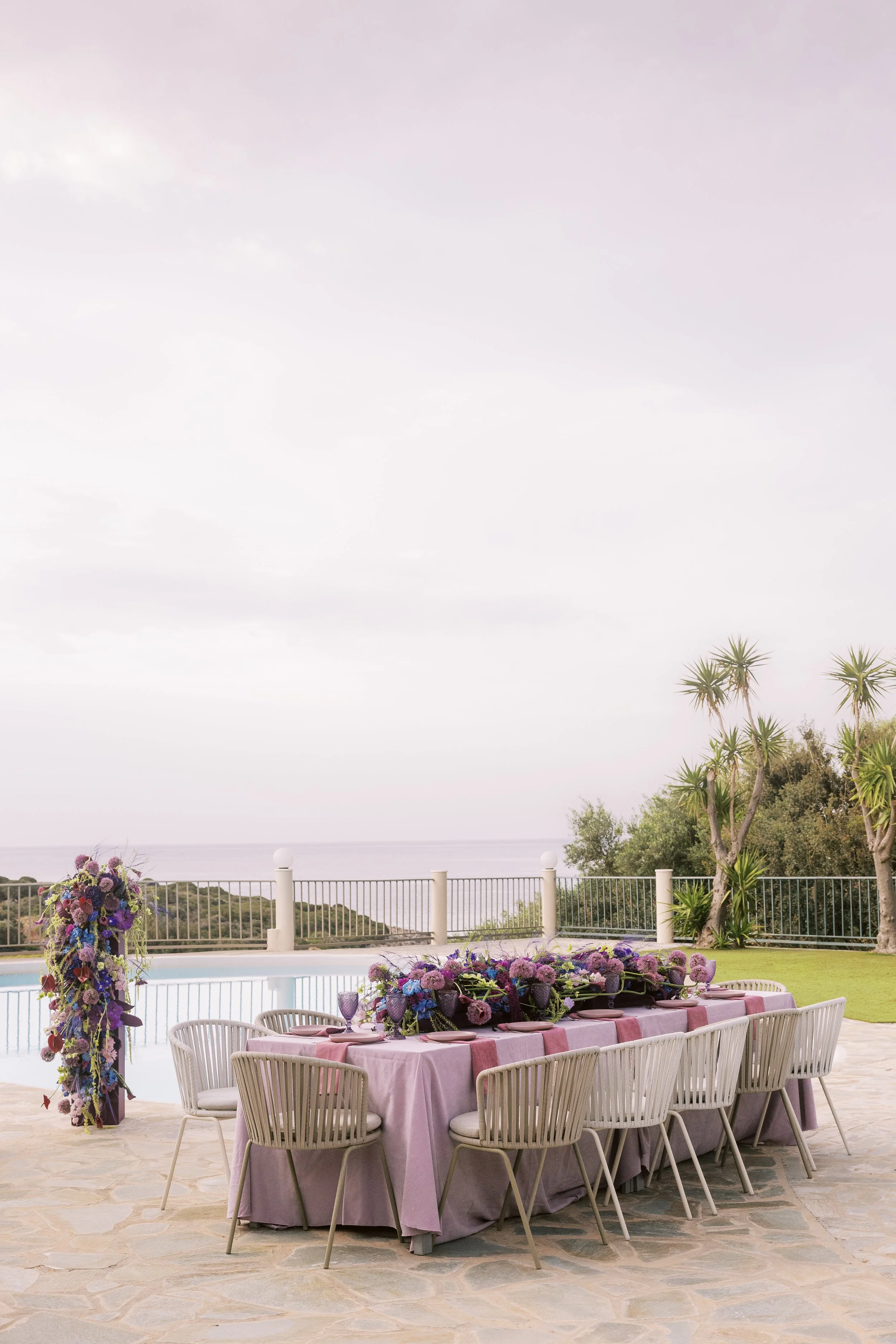 Outdoor event setup with a long dining table covered with a purple tablecloth, decorated with purple and pink flowers, and surrounded by white chairs. The setting overlooks a scenic view of the ocean with a cloudy sky, palm trees, and a white fence.