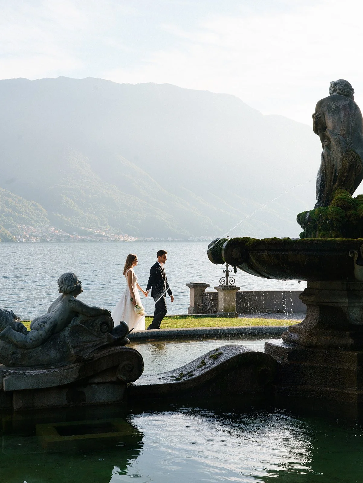 Bride and groom walking beside a stone fountain in the gardens overlooking Lake Como.