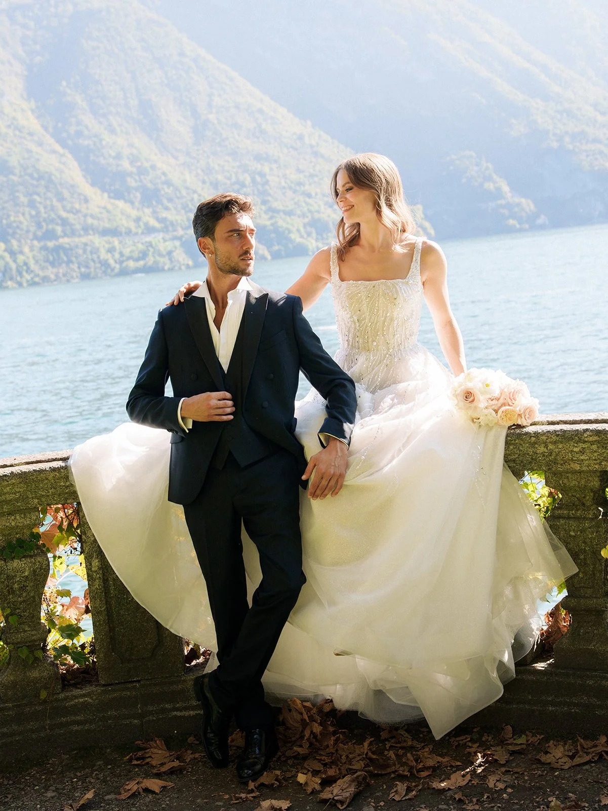 Bride and groom seated on a stone terrace overlooking Lake Como, with a pale pink bouquet beside the bride.