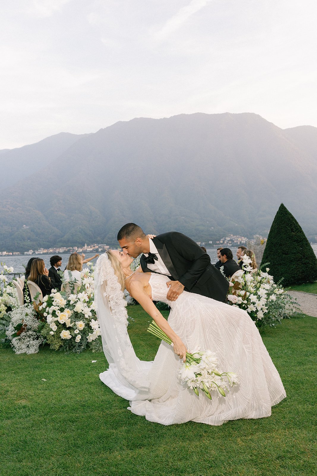 A couple dressed in wedding attire sharing a kiss during their outdoor wedding ceremony. The bride is in a white gown holding a bouquet of white flowers, and the groom is in a black tuxedo. There are guests seated behind them with a scenic mountainous lake view.