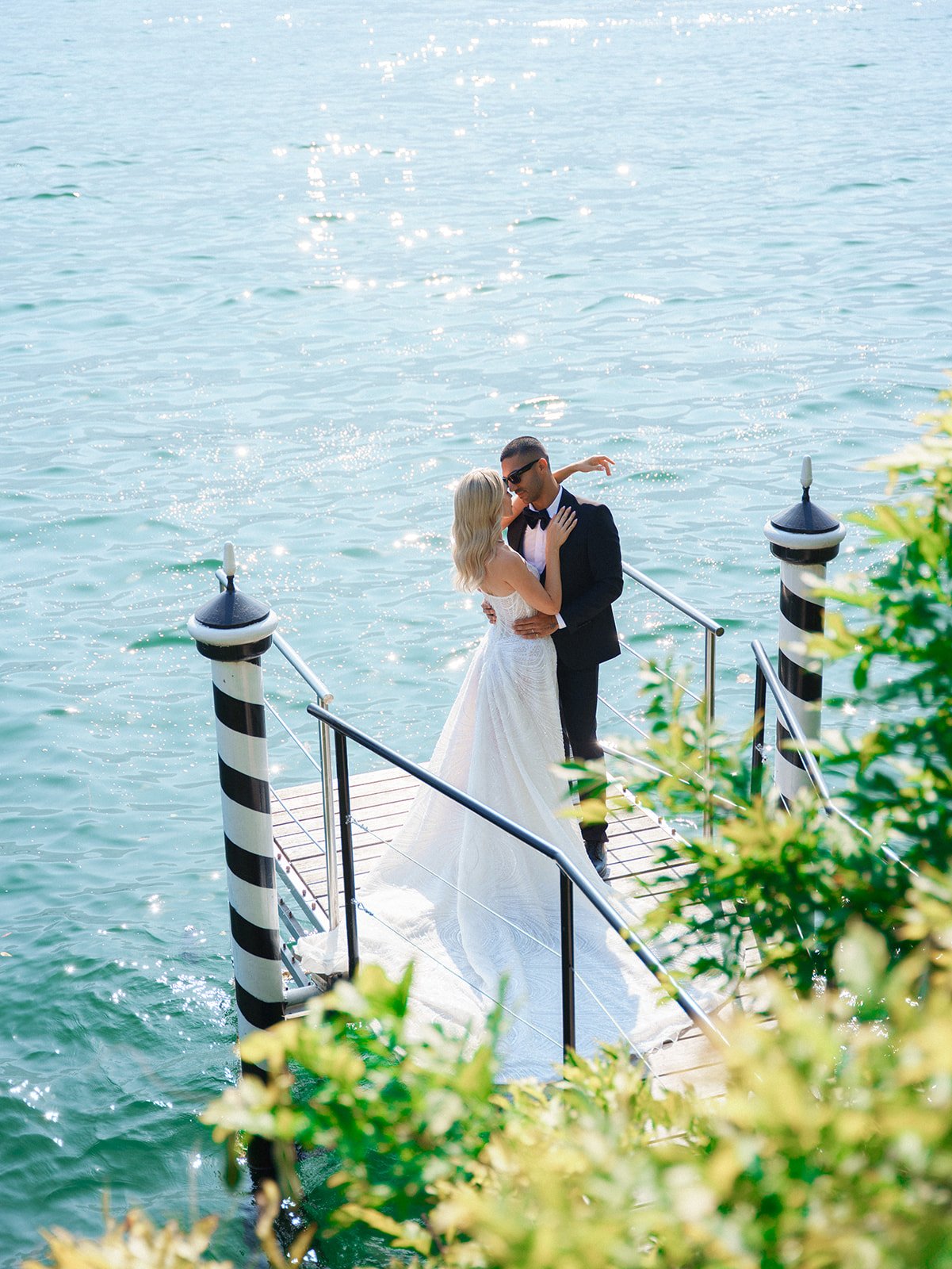 A bride and groom dancing on a dock over water, with the bride in a white wedding dress and the groom in a black tuxedo, surrounded by a railing with striped black and white posts, greenery partly in the foreground.