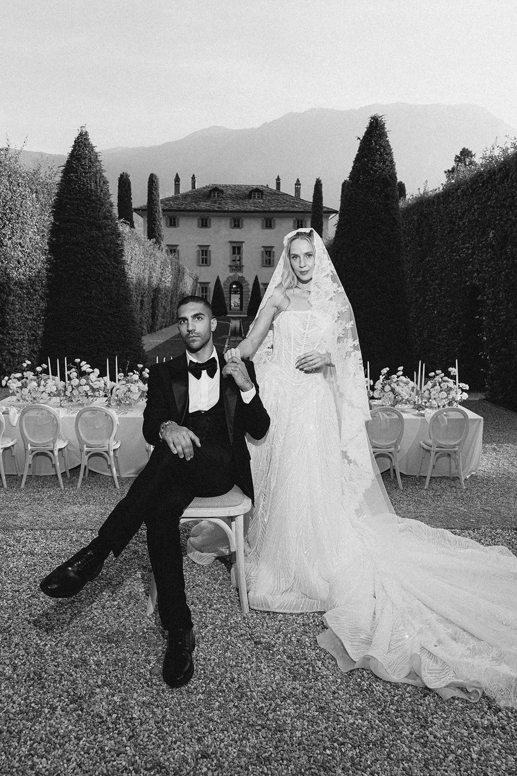 A black and white photo of a man in a tuxedo and a woman in a wedding dress standing outdoors in front of a grand house with a garden and mountain in the background.