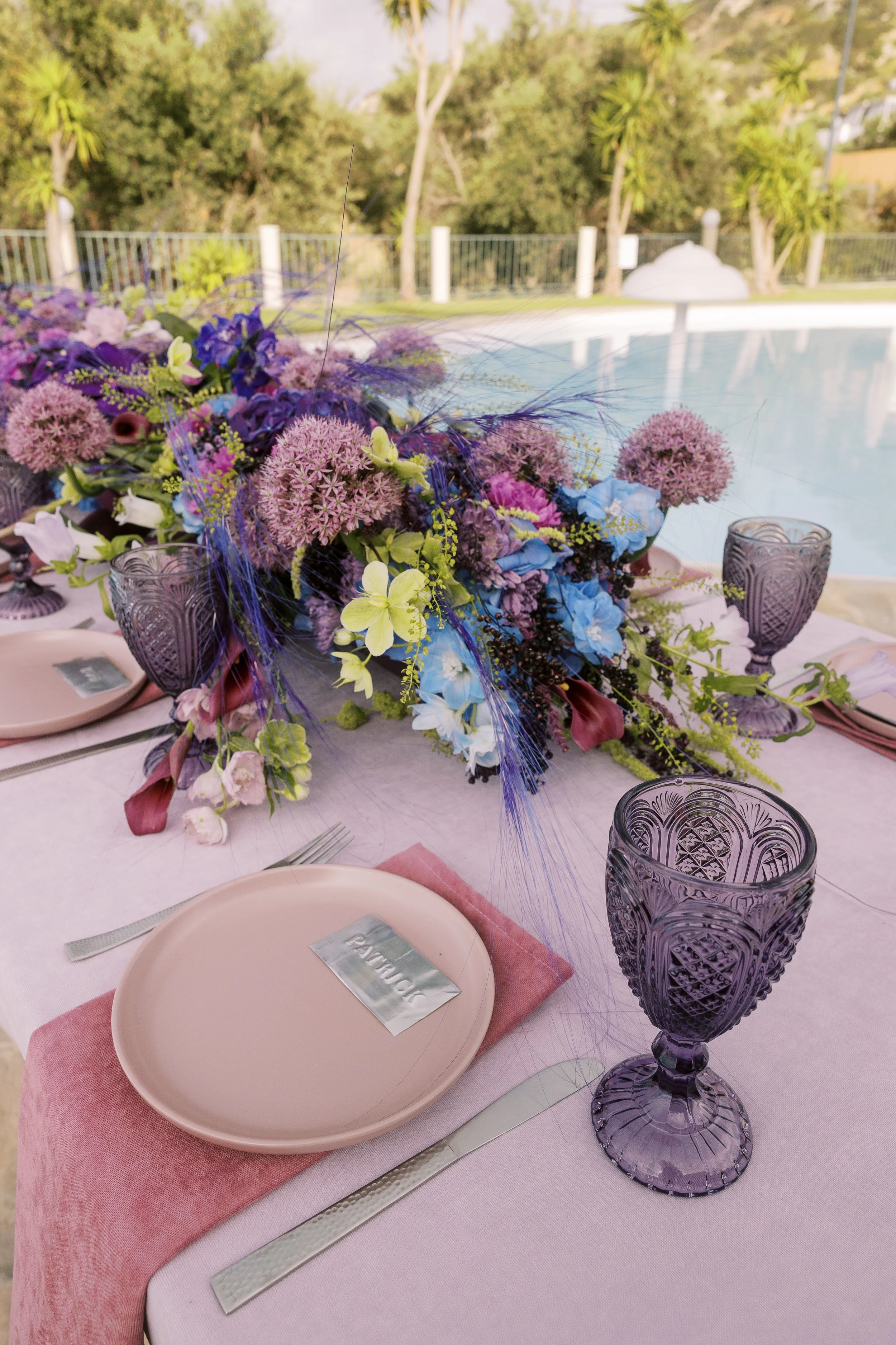 Smoky purple goblets beside layered violet florals and soft pink wedding tableware.