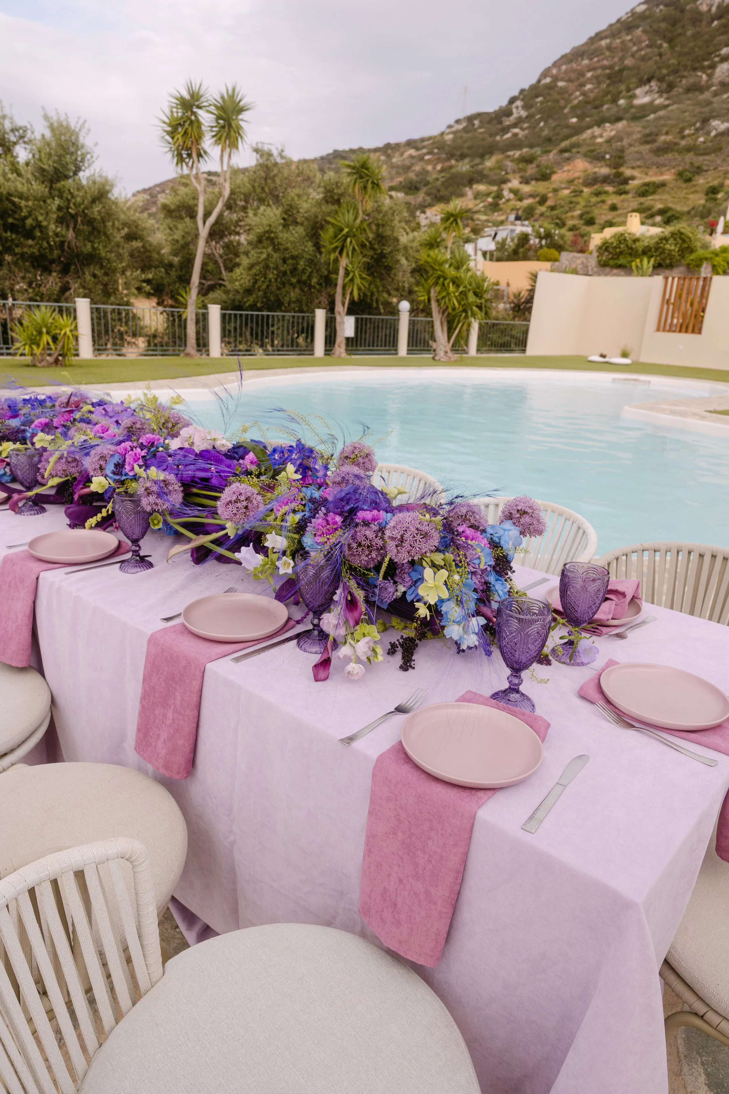 Colourful wedding reception table beside the pool with lilac linens, pink tableware and purple floral design.