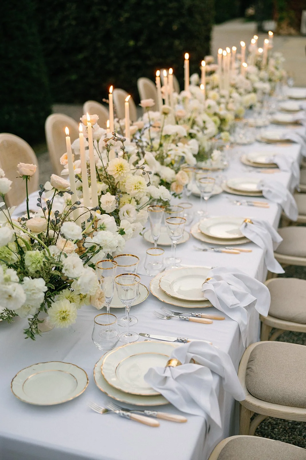 Elegant outdoor banquet table decorated with white flowers, tall candles, fine china with gold rims, crystal glasses, and whitenapkins, set for a wedding or special event.