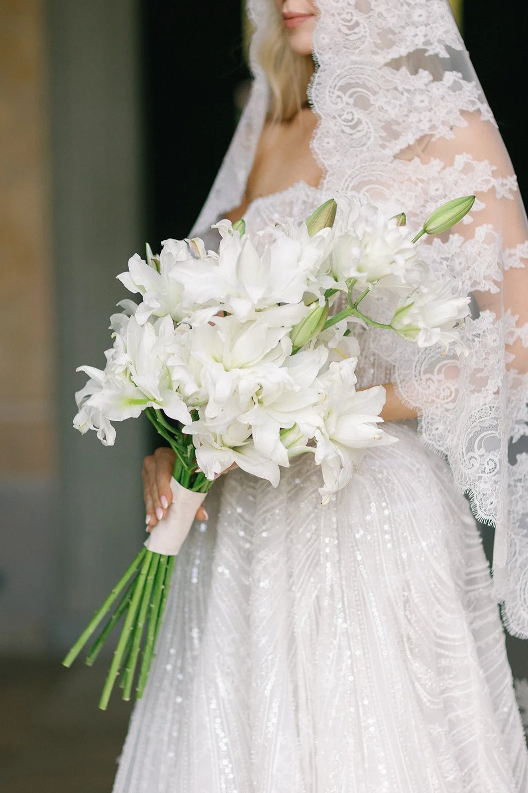 Close-up of a bride in a lace wedding gown holding a sculptural bouquet of white lilies.