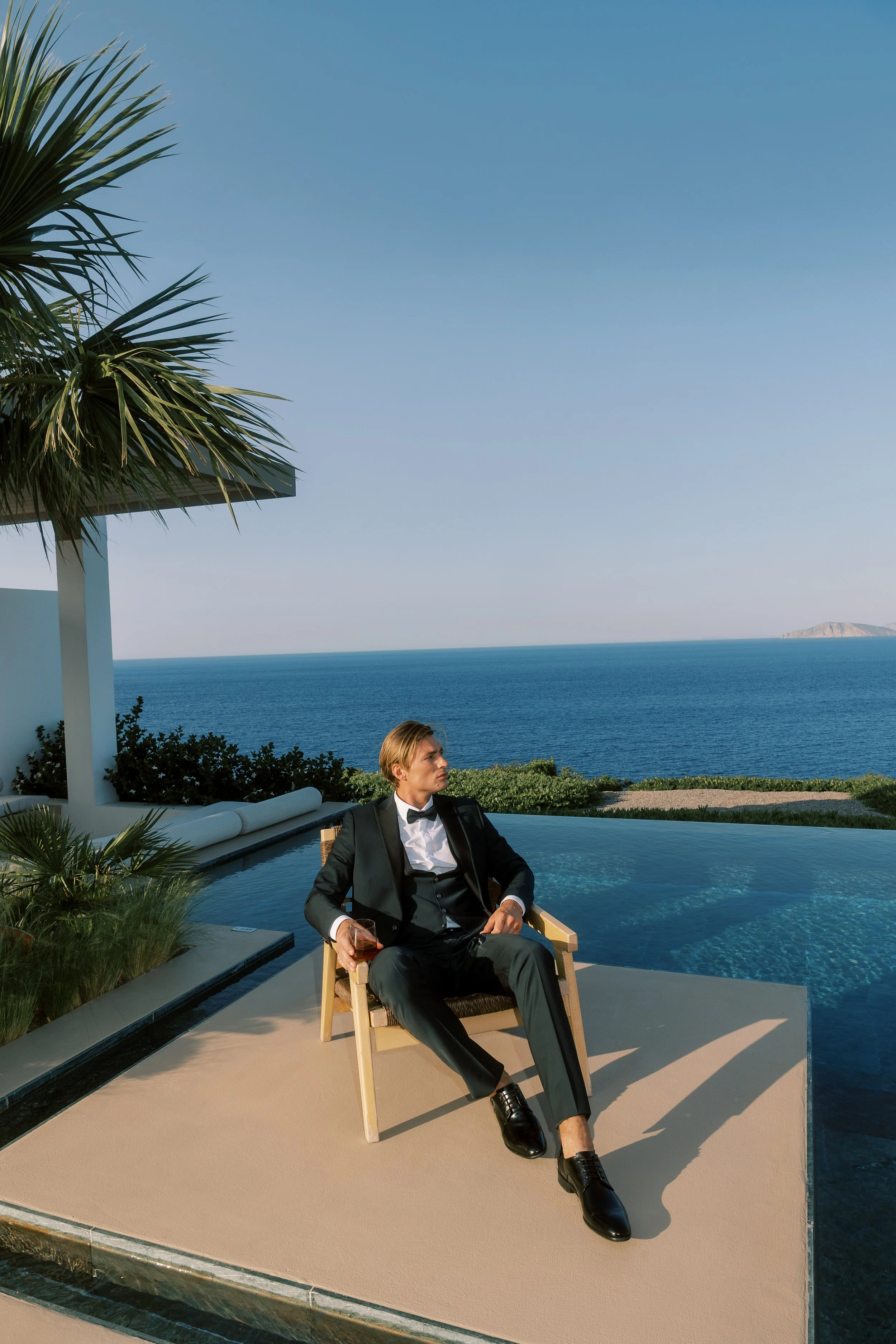 A man in formal attire sitting on a patio by a pool, holding a glass of wine, overlooking the ocean with a clear sky and distant land or island in the background.
