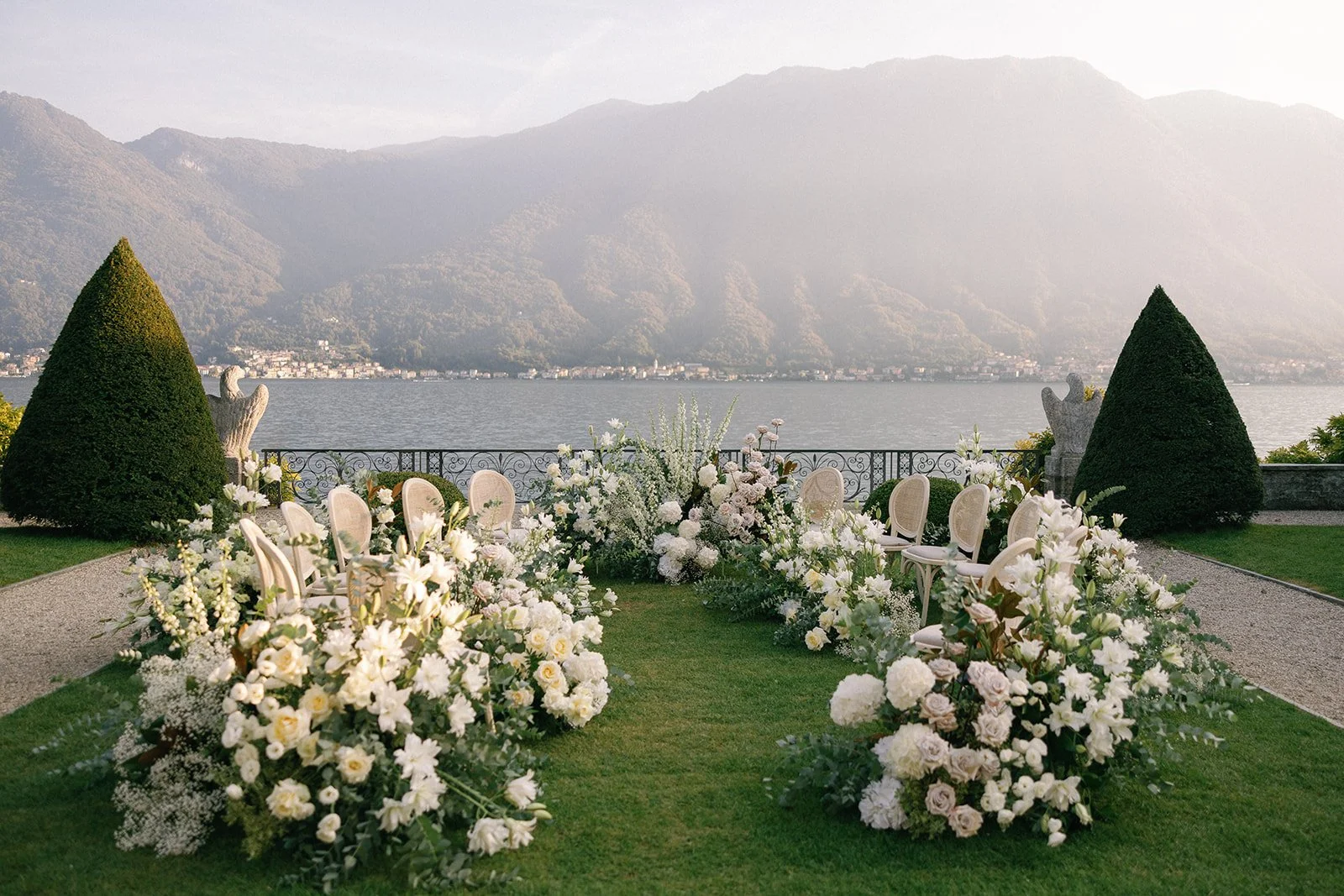 Elegant outdoor wedding setup with white flowers, chairs, and lush greenery by a lake with mountains in the background.