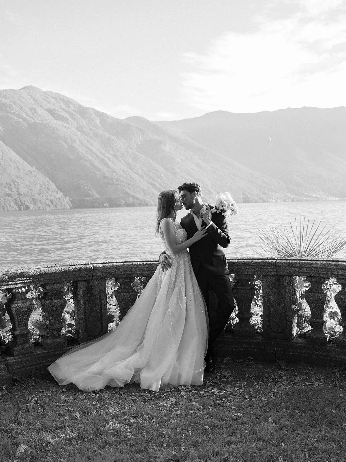 Black and white photo of a bride and groom embracing on a stone balcony overlooking a lake with mountains in the background. The bride is wearing a wedding gown, and the groom is in a suit holding a bouquet.