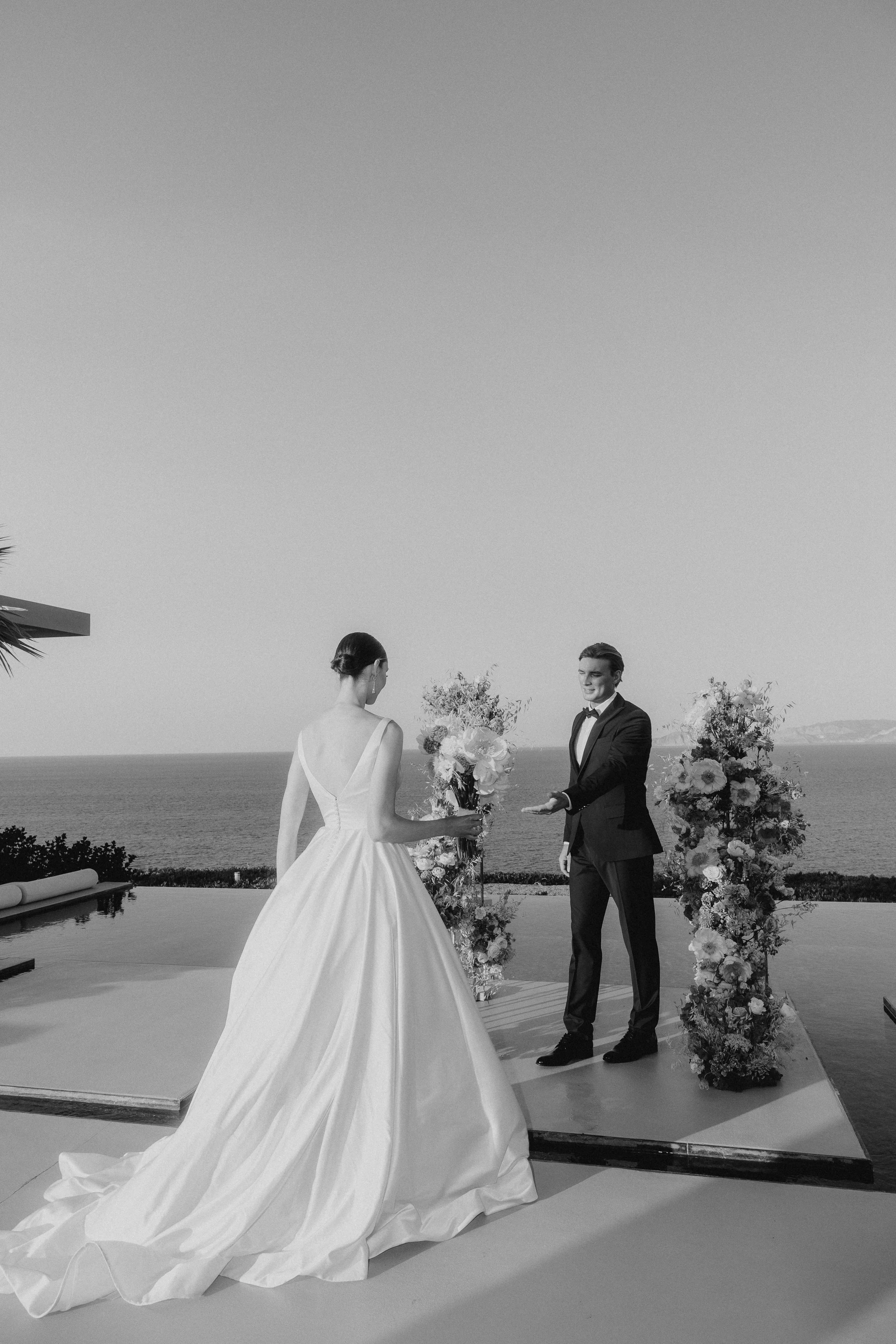 A bride and groom exchanging vows during a wedding ceremony outdoors with ocean and mountains in the background, decorated with floral arrangements.