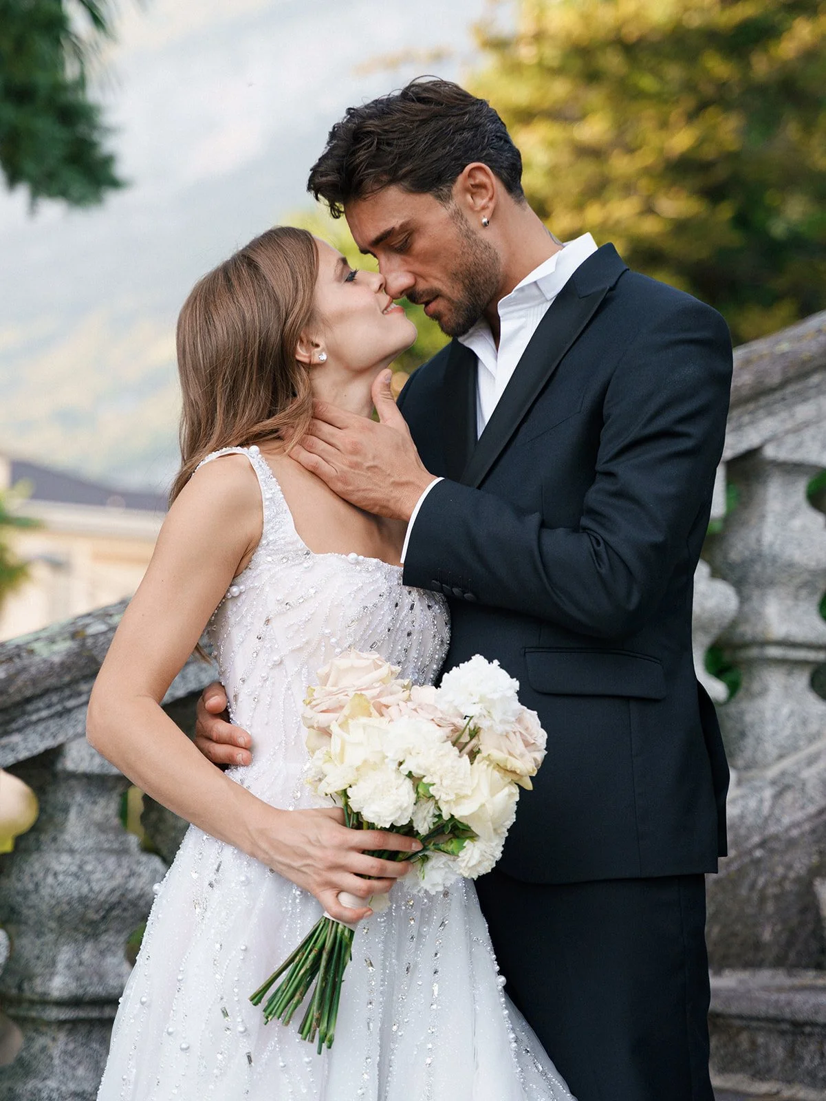A bride and groom close together, about to kiss, outdoors during daytime. The bride holds a bouquet of white and blush roses, wearing a white wedding dress, and the groom is in a black tuxedo with a white shirt.