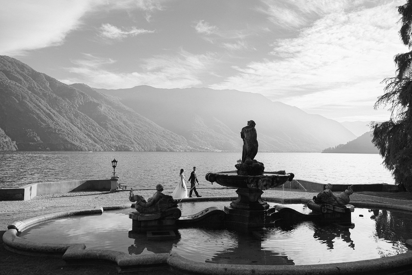 Black-and-white view of a couple walking through historic gardens beside a fountain overlooking Lake Como.