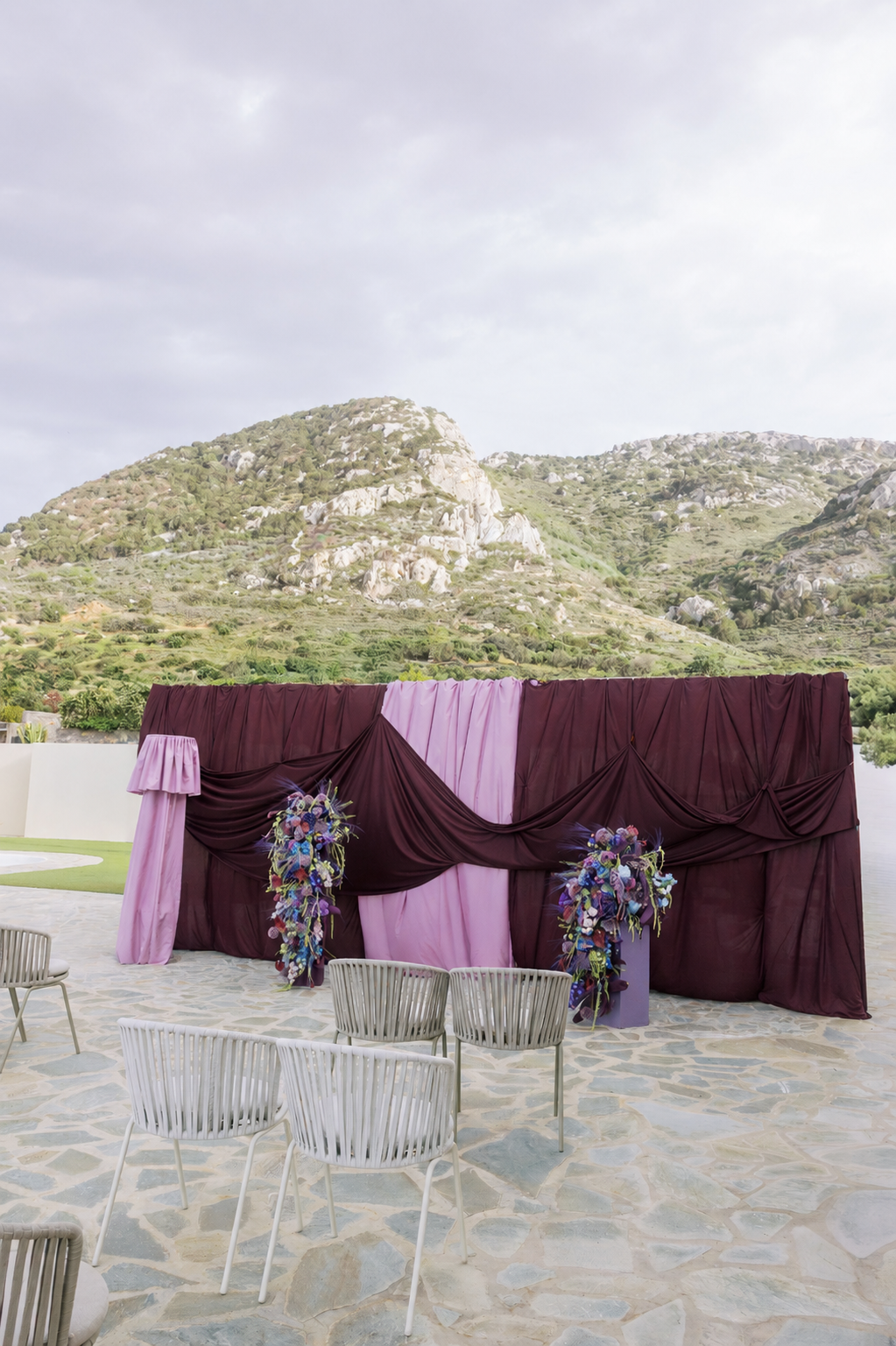 Colourful wedding tablescape in Crete with purple florals, lilac linens, smoky glassware and an aubergine draped ceremony backdrop.