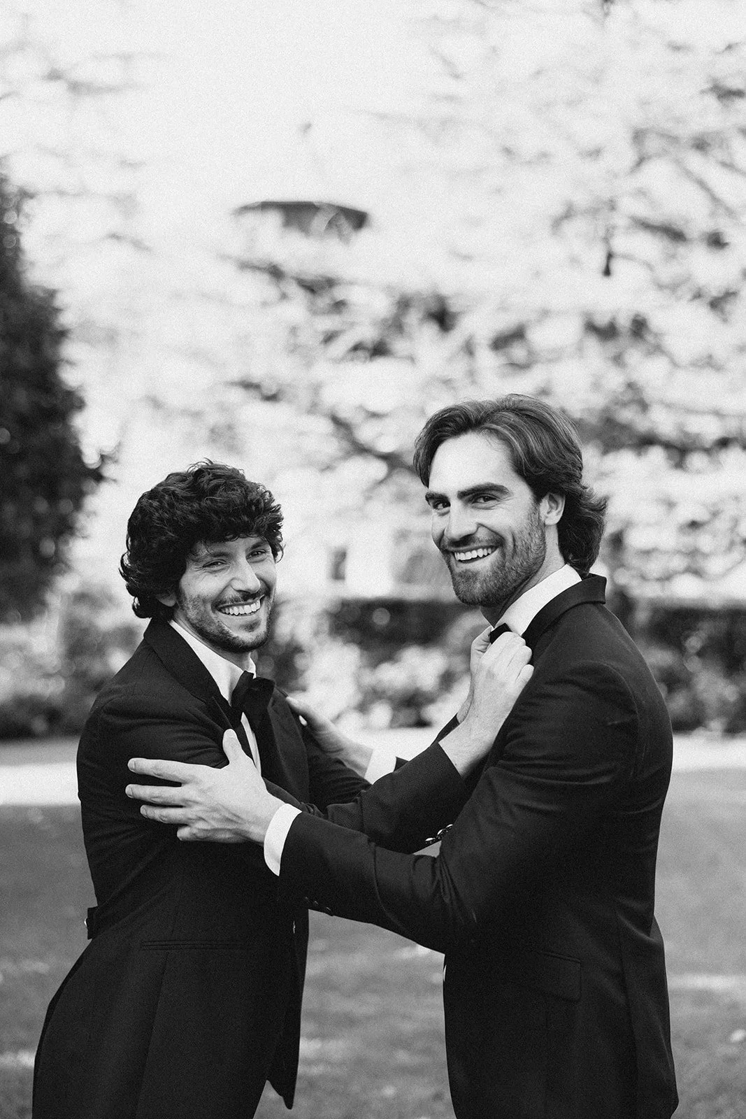 Two men in tuxedos smiling and holding each other in a friendly embrace outdoors, with trees and a building in the background.