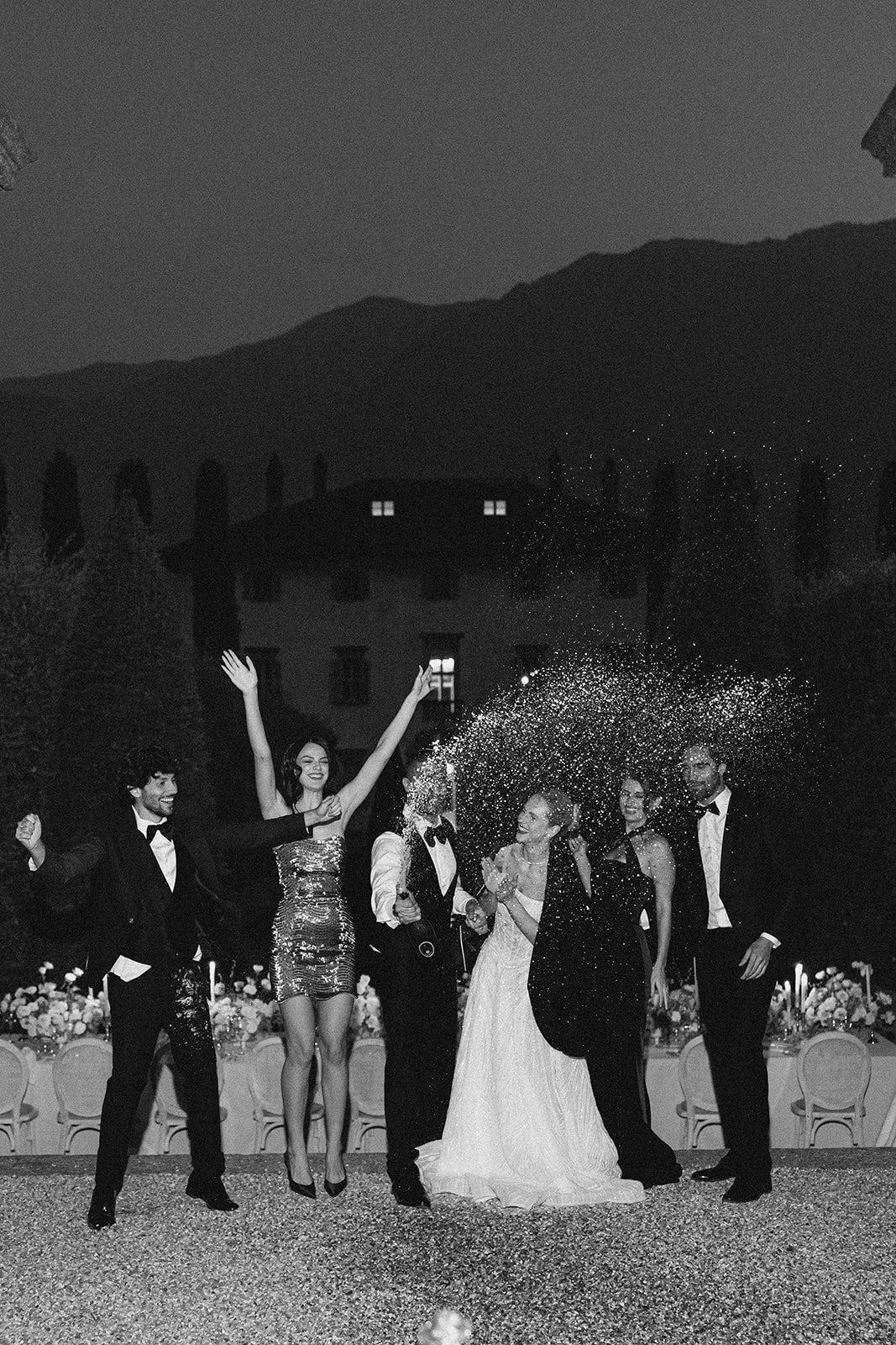 A group of people dressed in tuxedos and evening gowns celebrating at a wedding reception outside at night with mountains in the background. They are throwing confetti or glitter in the air, and there are tables with floral centerpieces behind them.