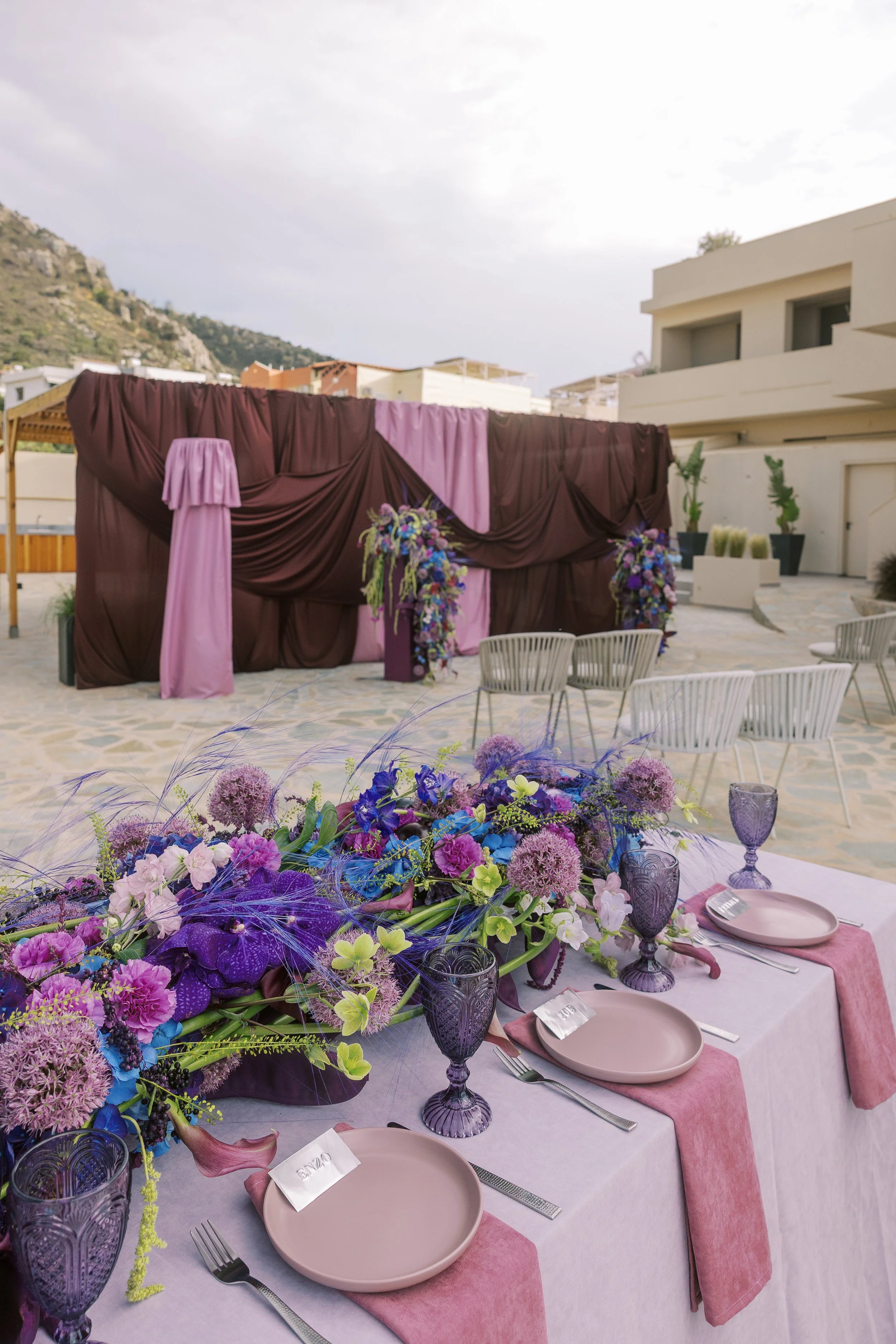 Colourful wedding tablescape in Crete with purple florals, lilac linens, smoky glassware and an aubergine draped ceremony backdrop.