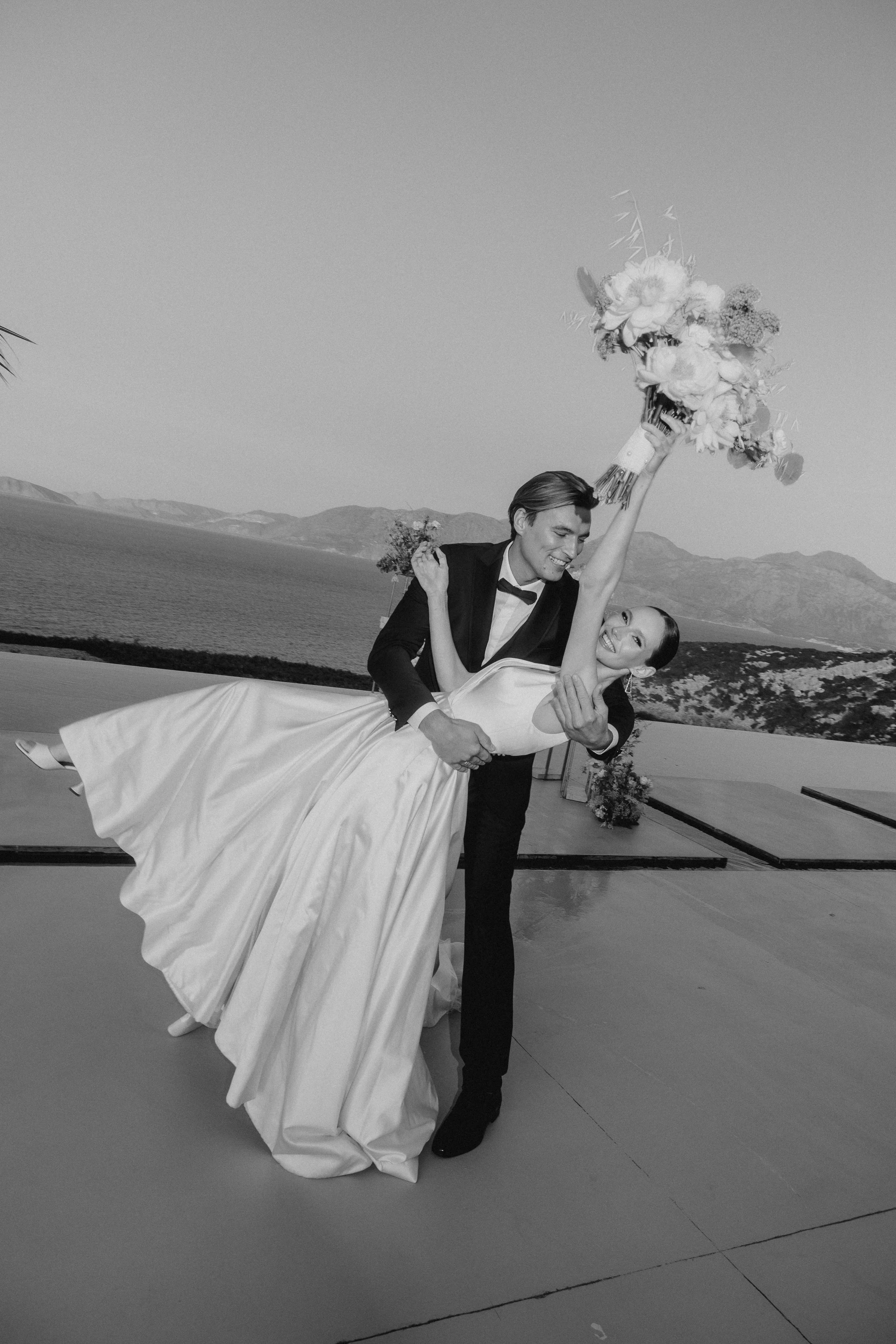 Black-and-white portrait of a groom dipping a bride holding a bouquet on a sea-view terrace in Crete, Greece.
