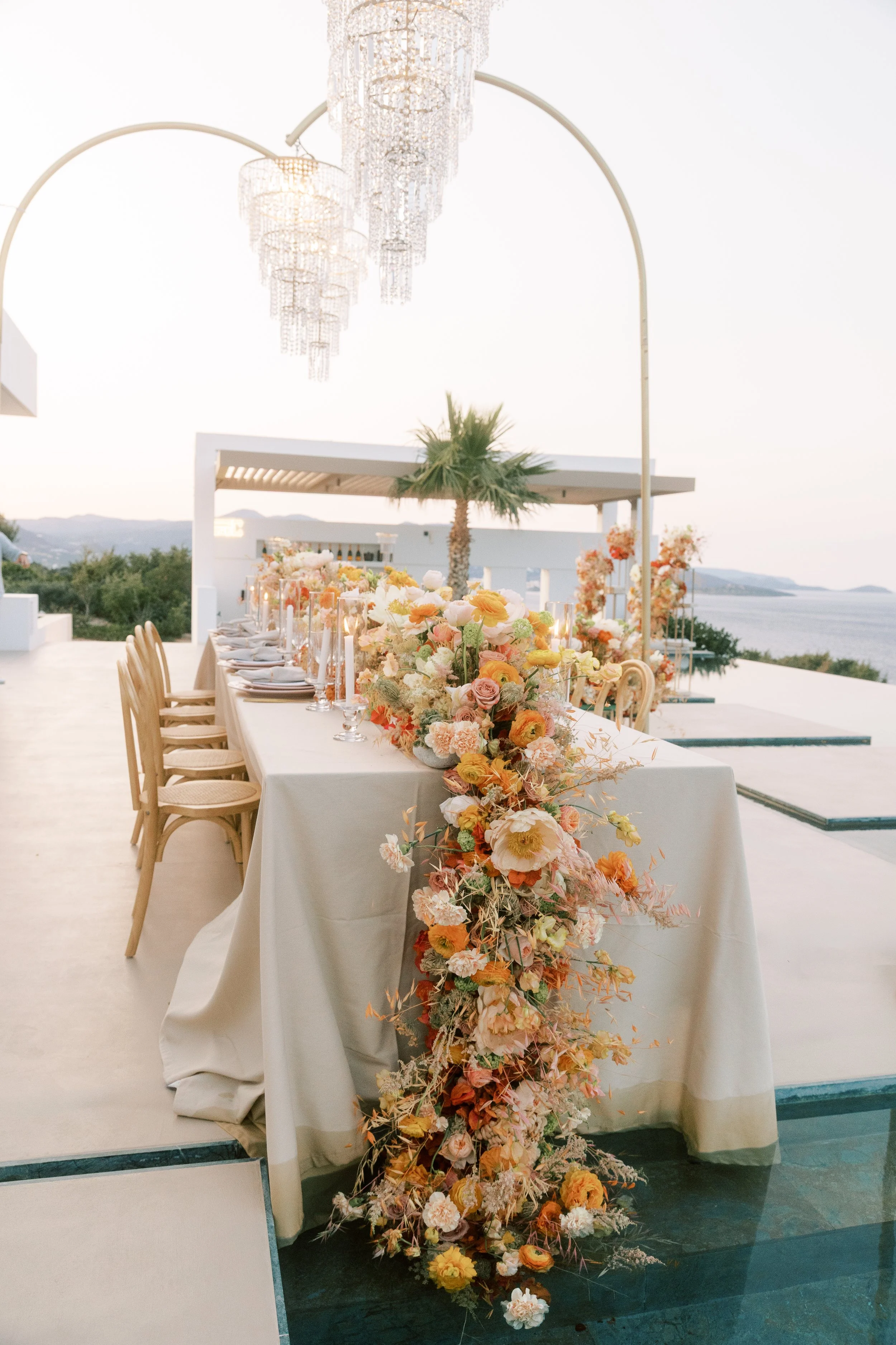 Long wedding table with a cascading floral runner under a crystal chandelier arch by the sea