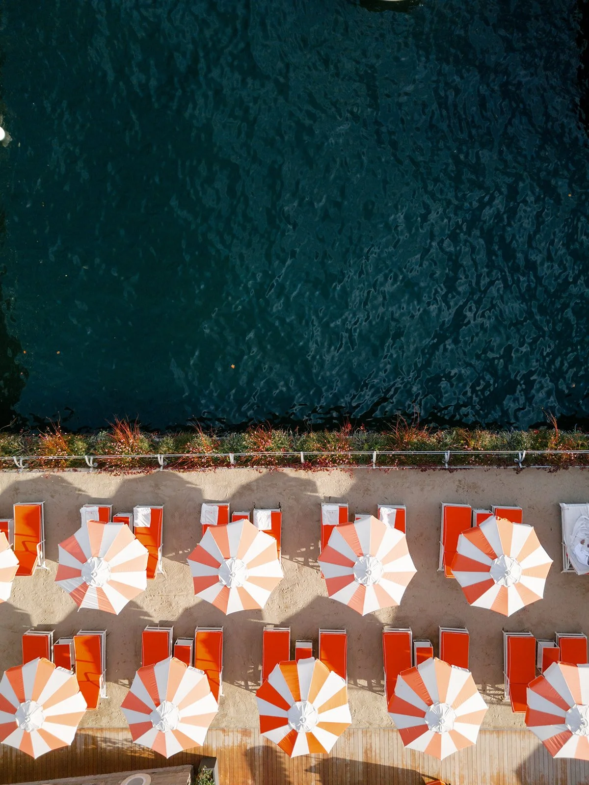 Aerial view of a beach area with orange and white striped umbrellas, lounge chairs, and a concrete border with plants along the water, which has a slightly rippled surface.