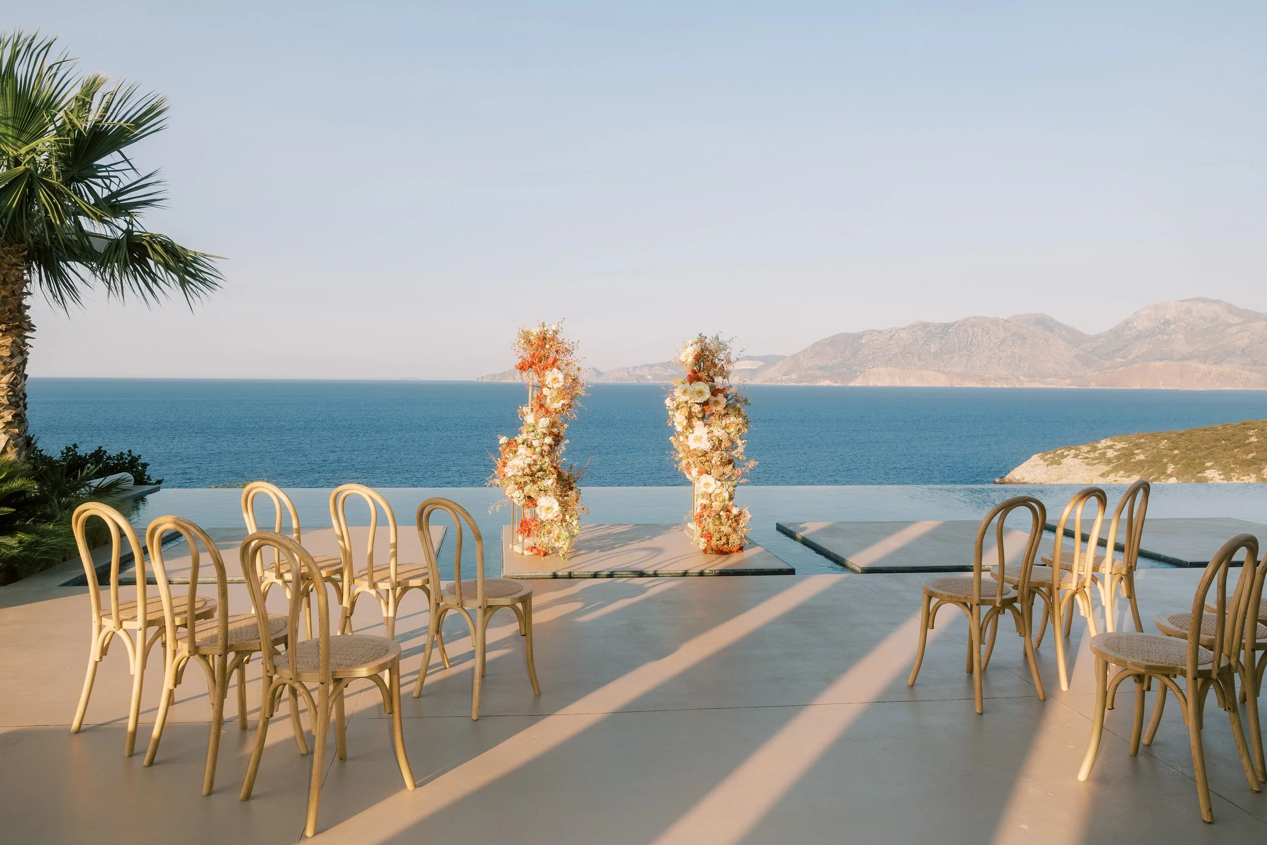 Outdoor wedding setup with chairs arranged around a floral decorated aisle, overlooking a large body of water with mountains in the background, and a palm tree on the left side.