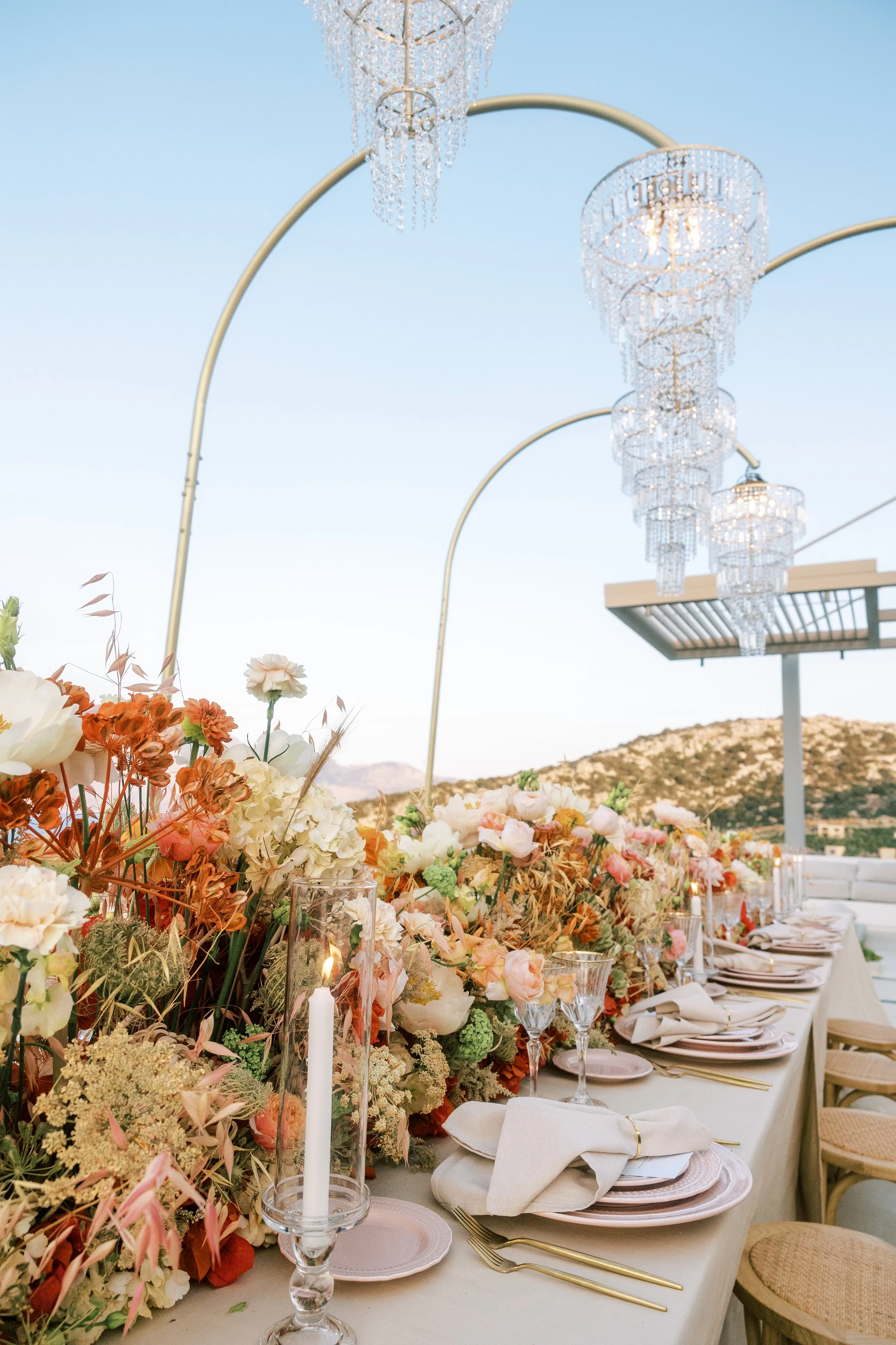 Elegant outdoor table setting with floral centerpieces, candlelit glass holders, and crystal chandeliers hanging above against a backdrop of blue sky and distant mountains.