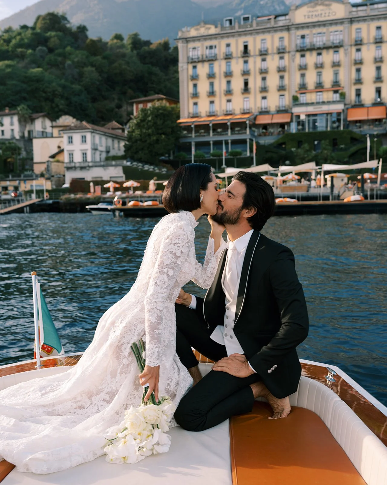 A newlywed couple sharing a passionate kiss on a boat in a scenic lakeside town with colorful buildings and lush green mountains in the background.