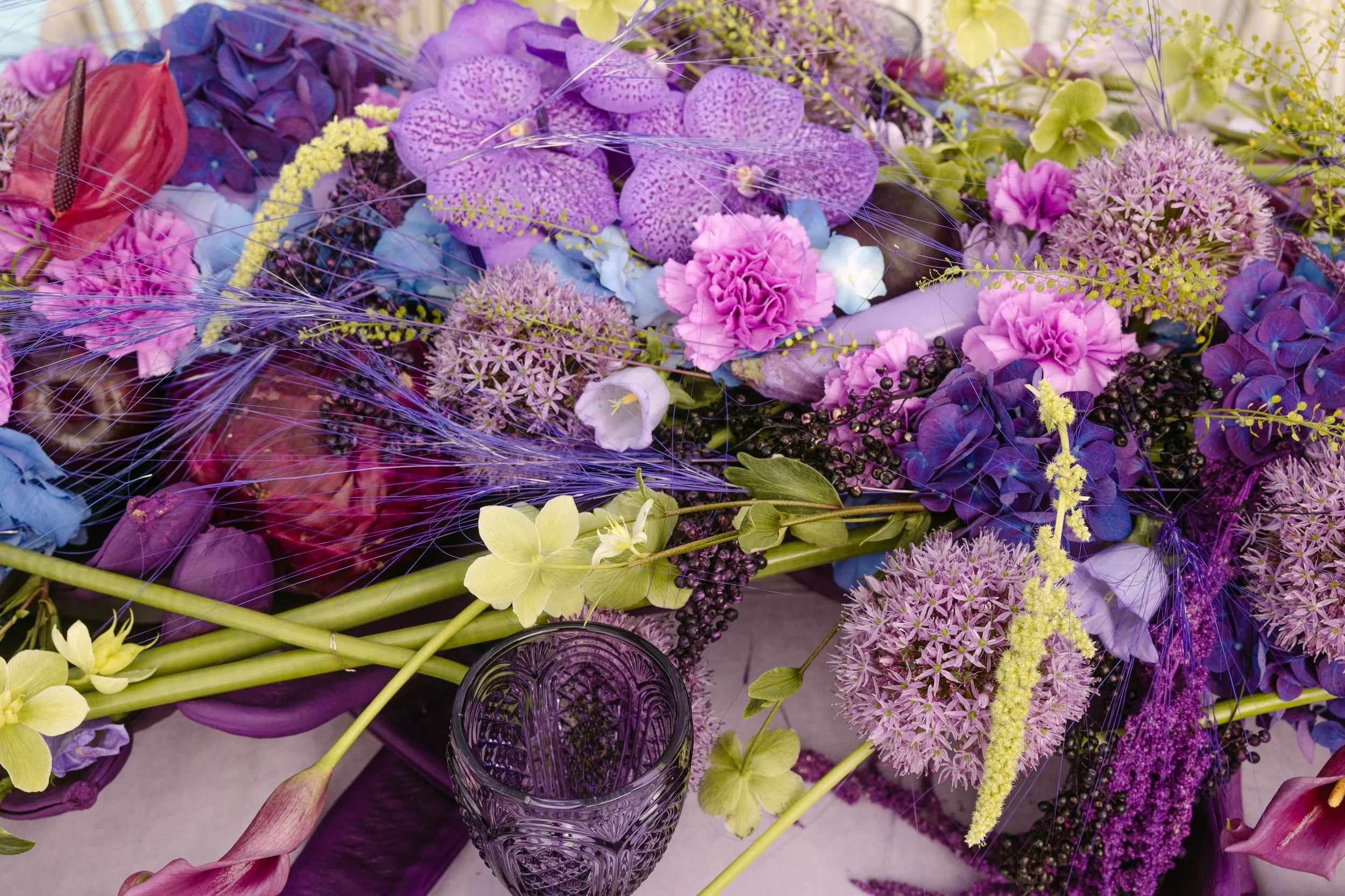 A close-up of a vibrant flower arrangement featuring purple, pink, yellow, and blue flowers, with a purple glass and a patterned glass visible in the scene.